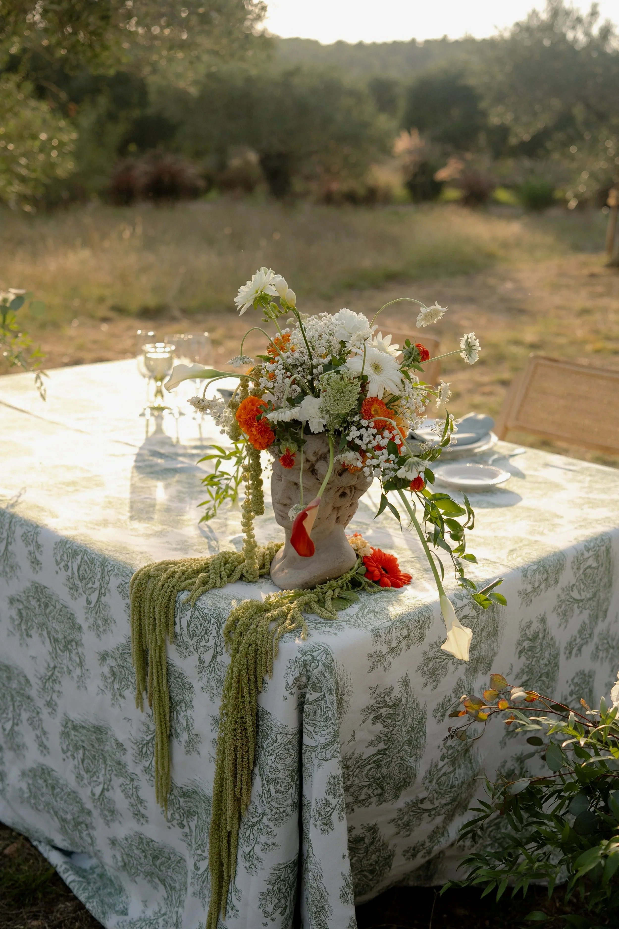 A table with a floral centerpiece of a rustical head, outdoors in a natural setting, decorated with white and orange flowers, green foliage, and a green garland, on a white and green english patterned tablecloth.