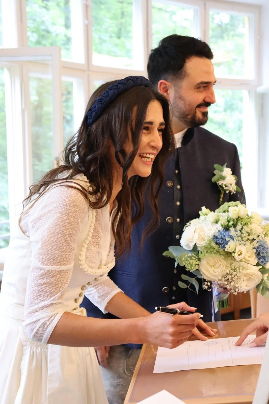 A woman in a white dress signs a document, smiling, holding a pen, with a man standing beside her holding a bouquet of white and blue flowers. They are inside a bright room with large windows and greenery outside.