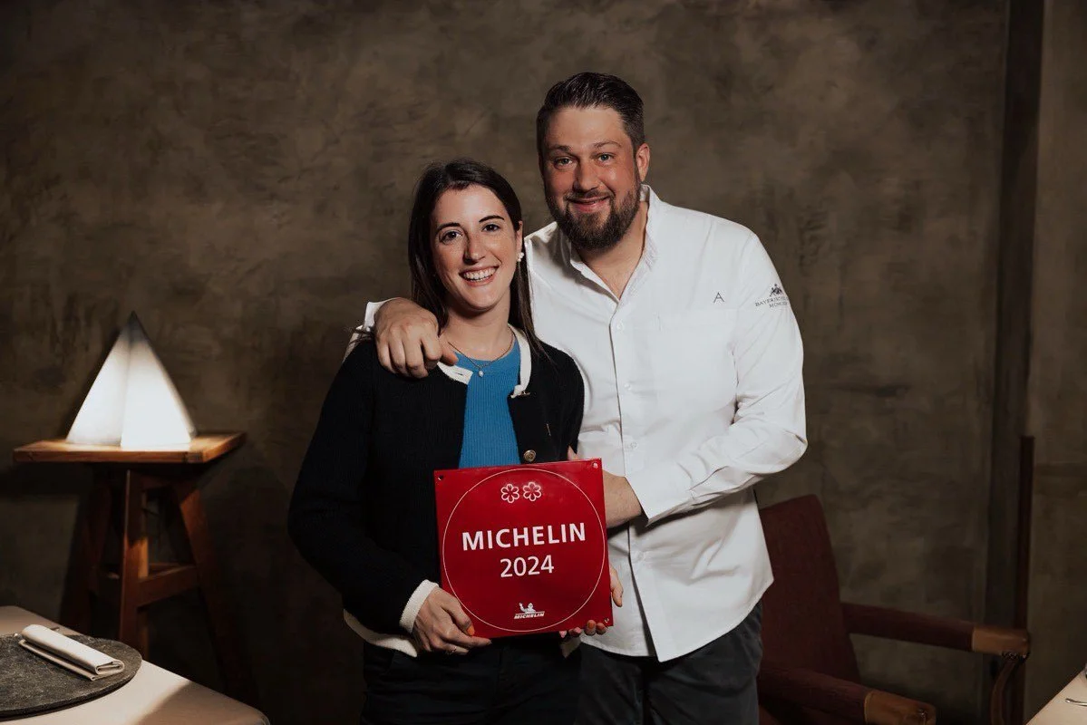 A smiling woman holding a red Michelin 2024 award plaque, standing next to a man with his arm around her shoulder in a restaurant setting.
