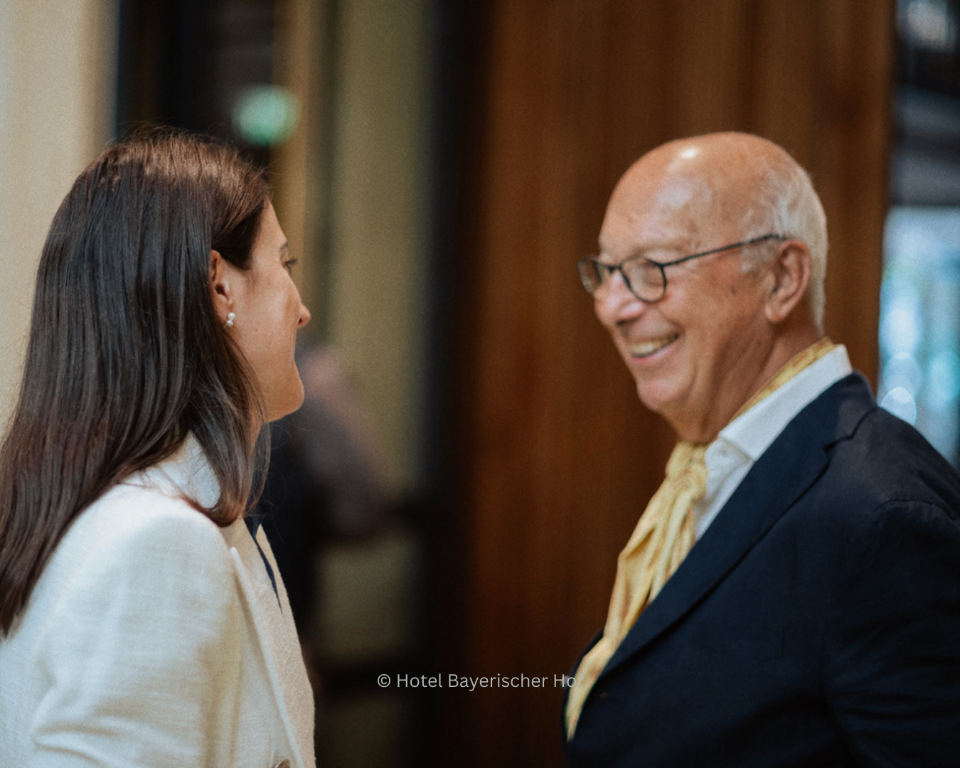 Two people, a woman and a man, are standing and smiling at each other in an indoor setting.