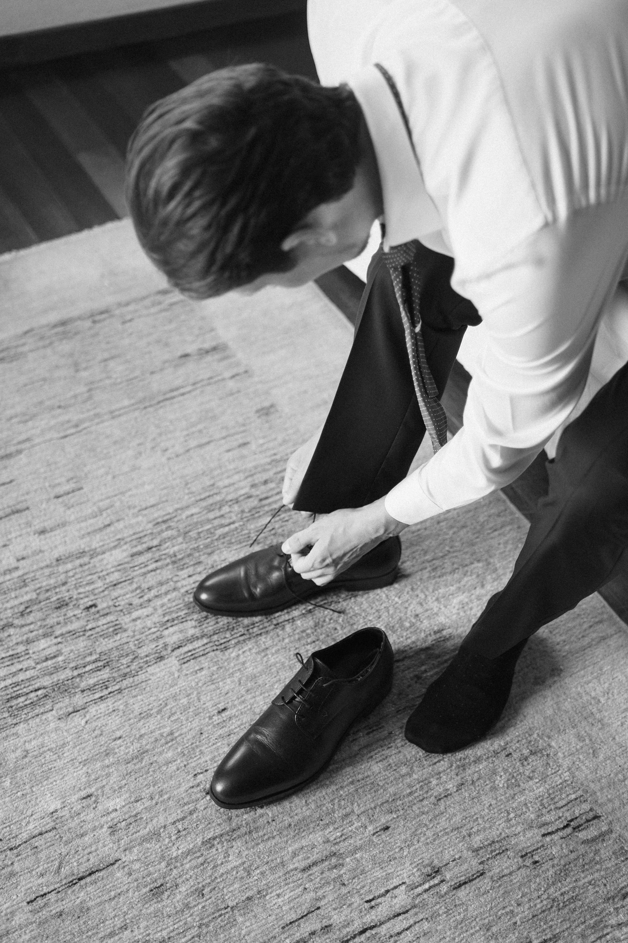 A man in formal attire, including dress shoes and socks, is bending over to tie his shoes while standing on a carpeted floor.