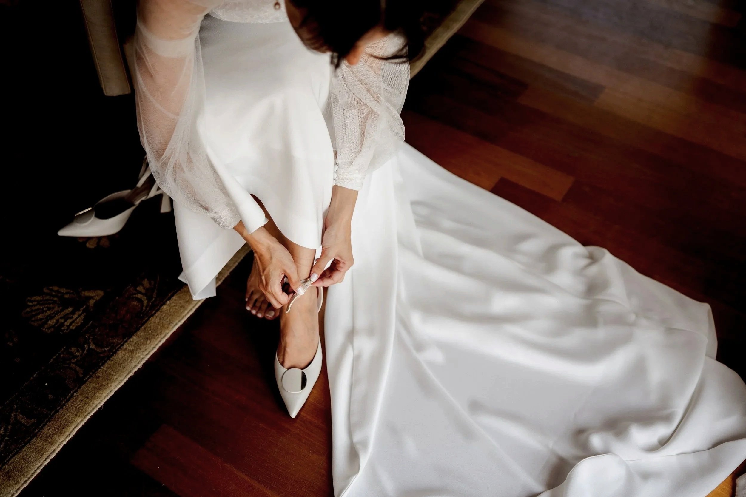 A bride in a white wedding dress sitting on a wooden floor, adjusting her white high heel shoe.