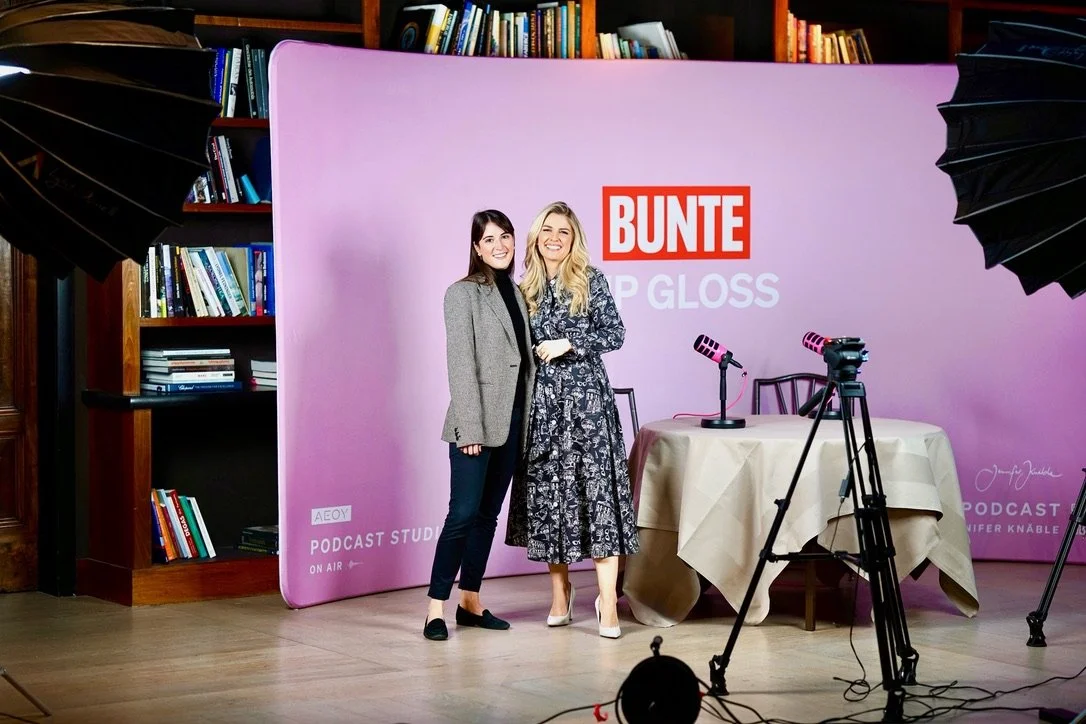 Two women standing in front of a purple backdrop with the words "BUNTE Lip Gloss" and podcast-related equipment, including a table with microphones, in a studio with bookshelves.