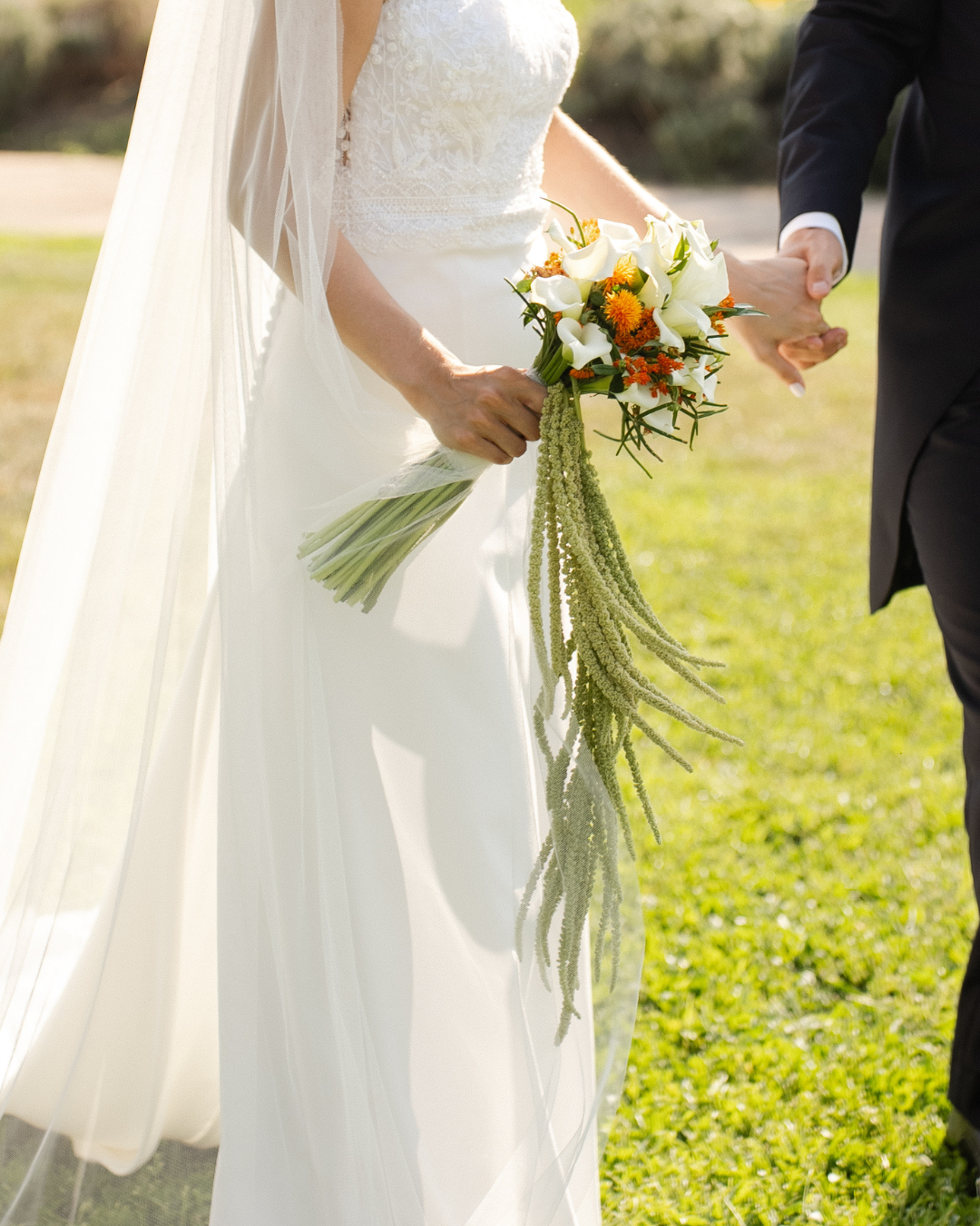 Bride holding a bouquet of white calla lilies, orange and yellow flowers, and green hanging foliage, during a wedding ceremony.
