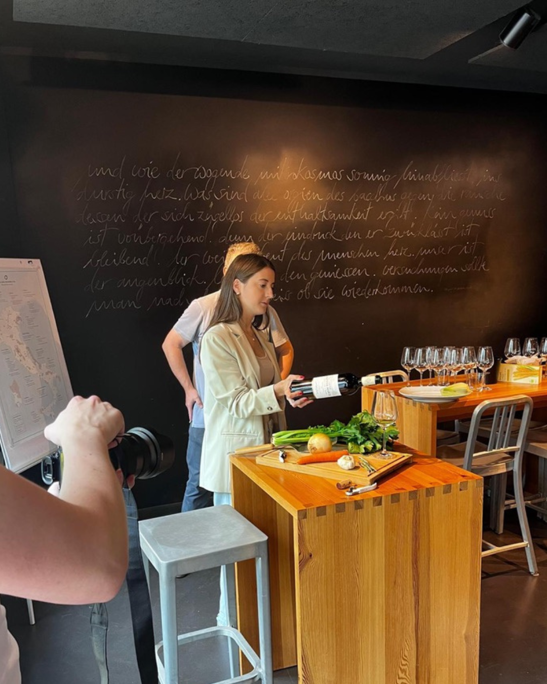 Woman holding a bottle of wine, preparing for a cooking or wine-tasting event in a modern kitchen or restaurant with vegetables and wine glasses on the counter.