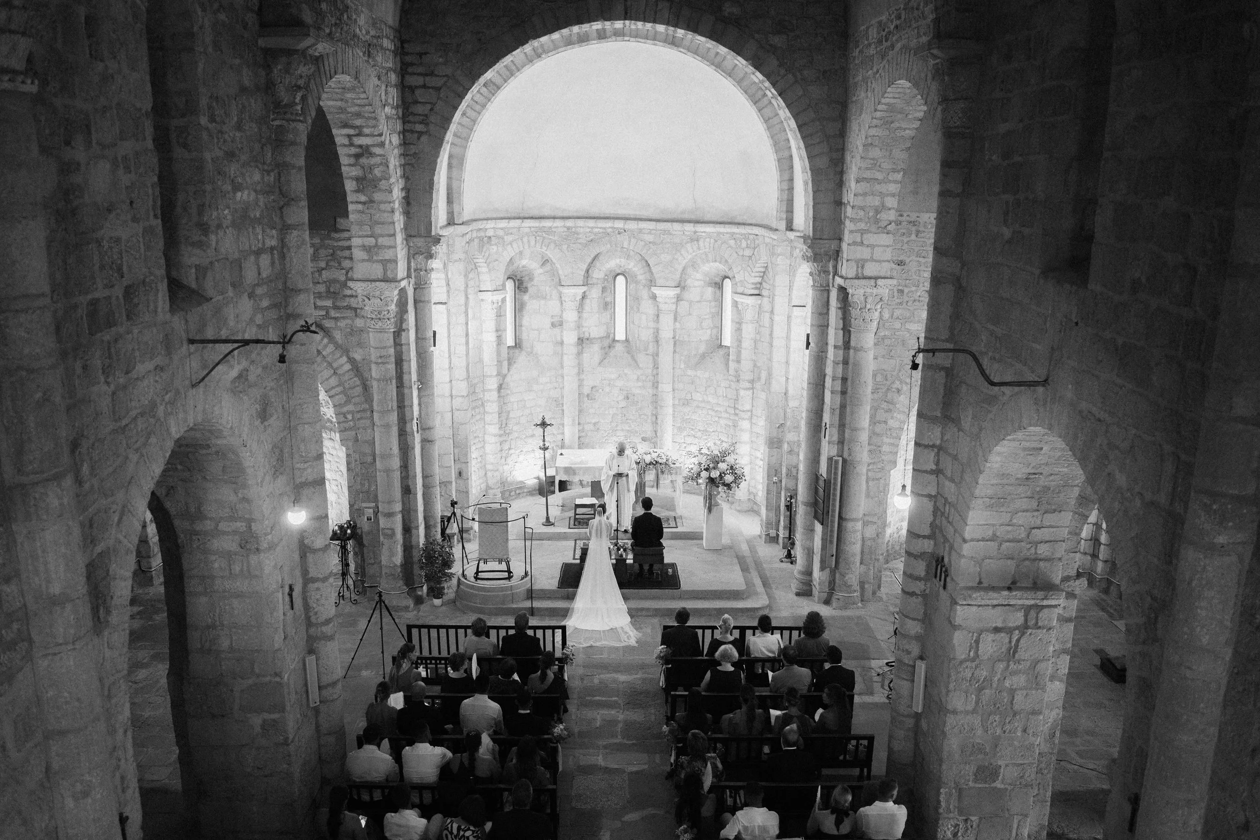 A black and white photo of a wedding ceremony inside the church of Sant Miquel de Fluvià in Spain with stone arches and columns, guests seated in pews, a priest officiating at the altar, and the bride and groom standing before him.
