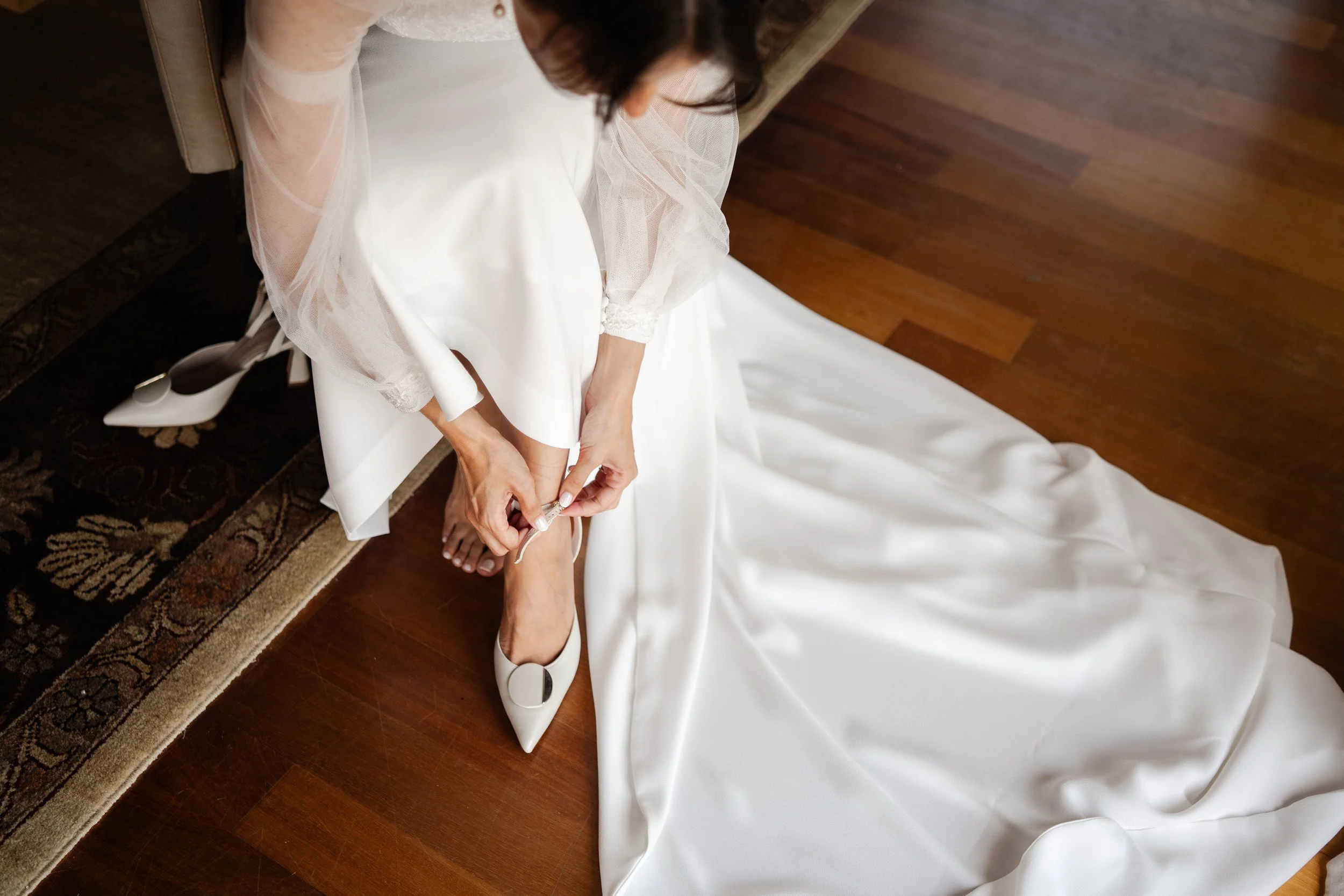 A bride in a white wedding dress is sitting on a chair, adjusting her white high-heeled shoe in a spanish wedding organised by Blanc Creative Studio.