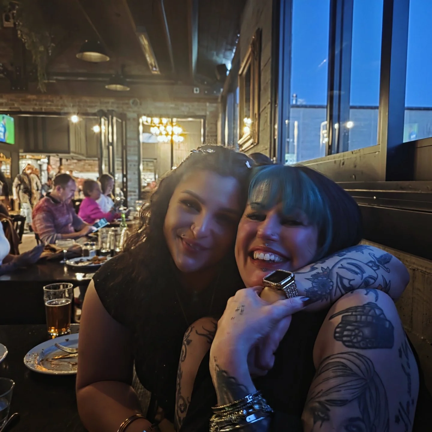 Two smiling women with tattoos hugging each other in a restaurant or bar, with people dining in the background and large windows showing a blue evening sky.