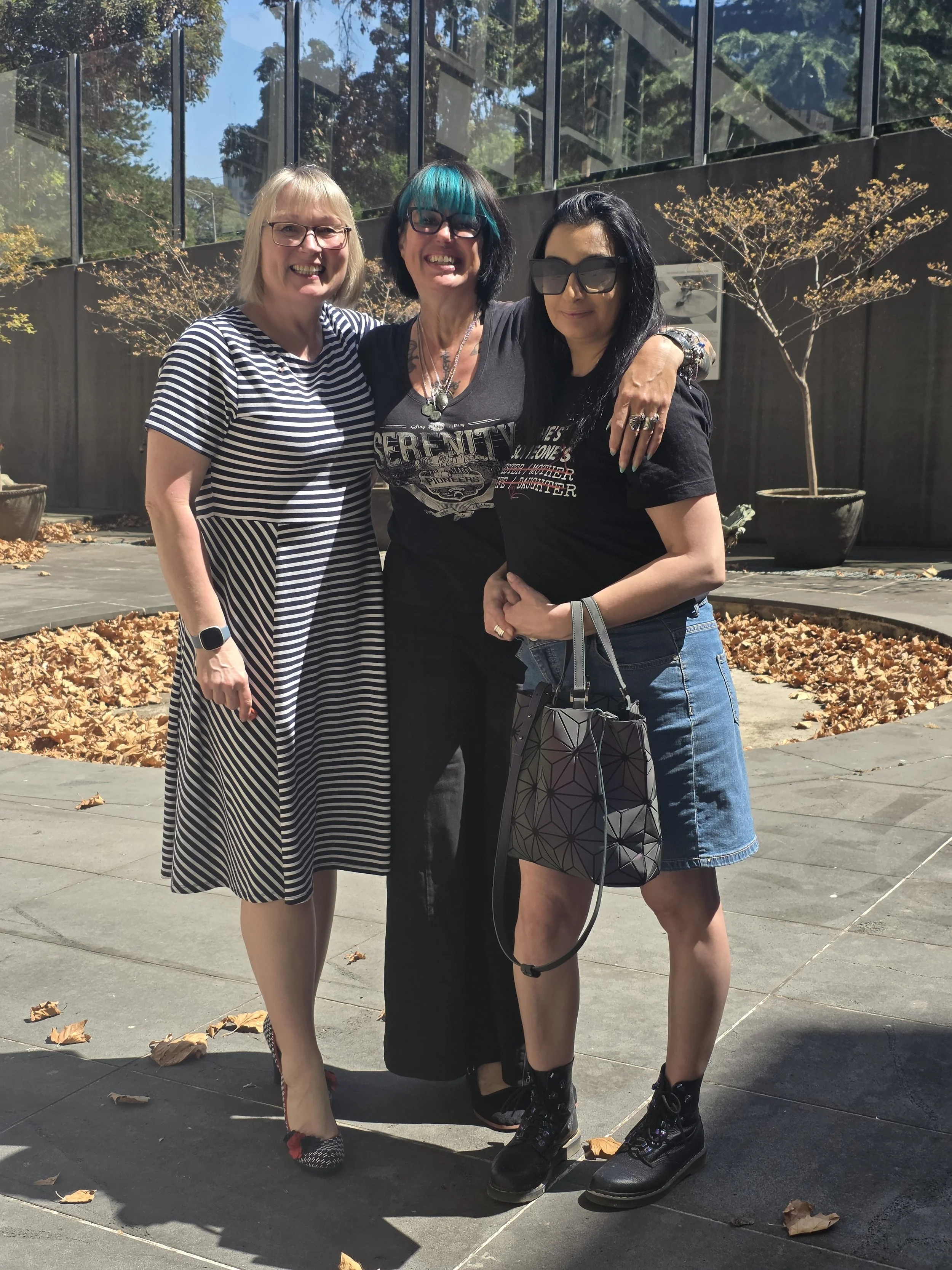 Three women standing outdoors on a sidewalk, smiling and posing for the photo with trees and a building with glass windows in the background.