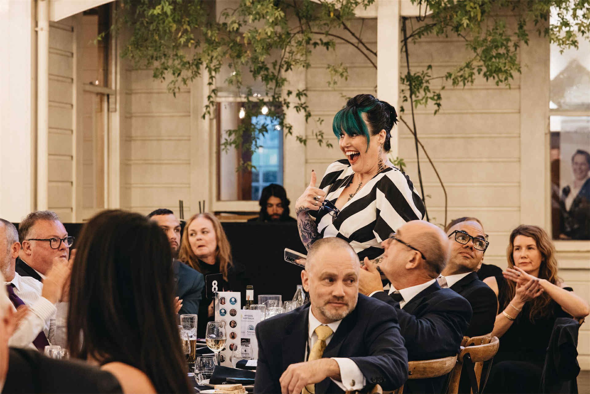 A woman with teal hair and tattoos, wearing a black and white striped dress, is standing and laughing, giving a thumbs-up, at a formal dinner event.