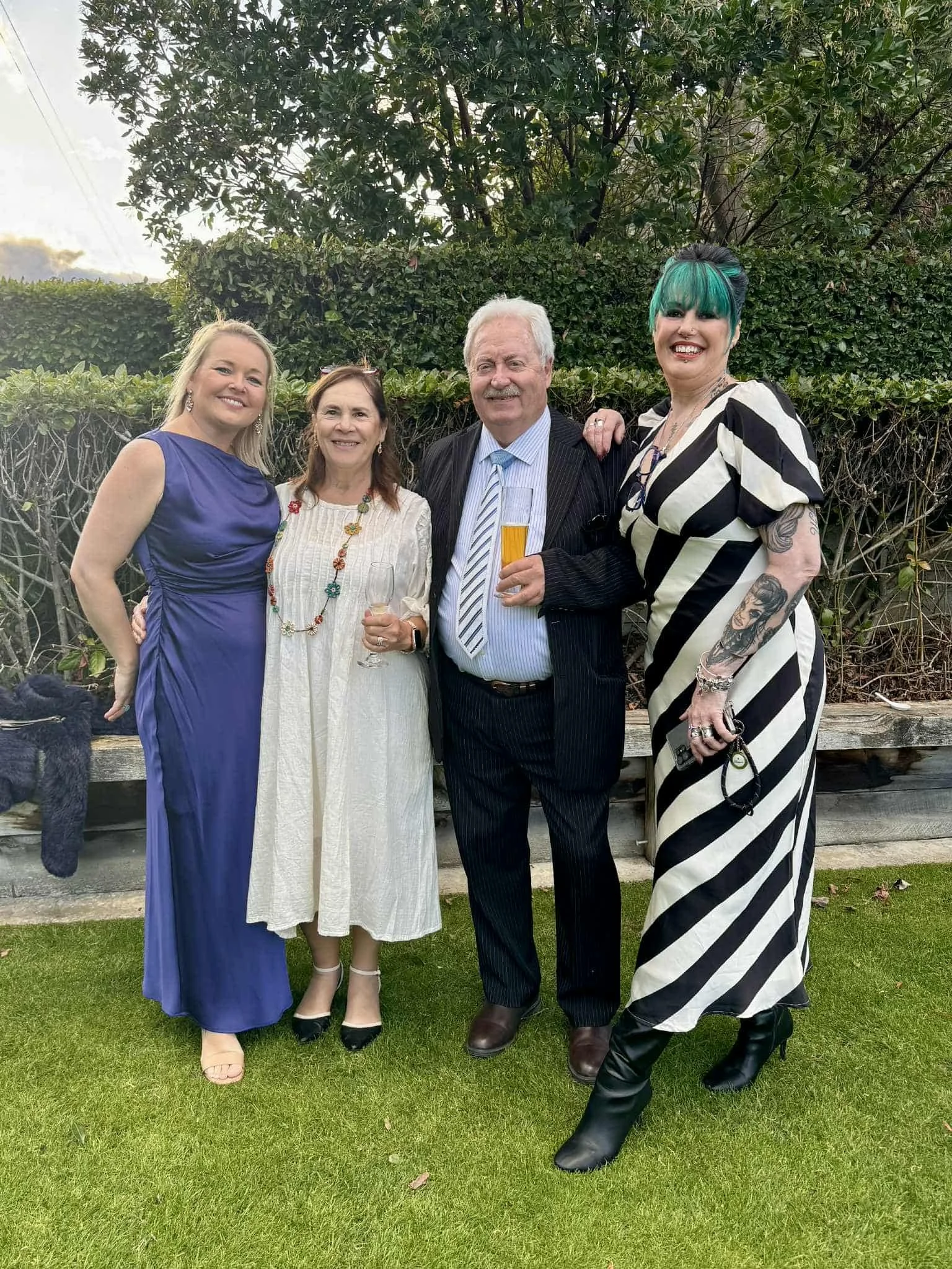 Group of four people dressed formally, standing outside on the grass, holding drinks, smiling for the camera with a background of bushes and trees.