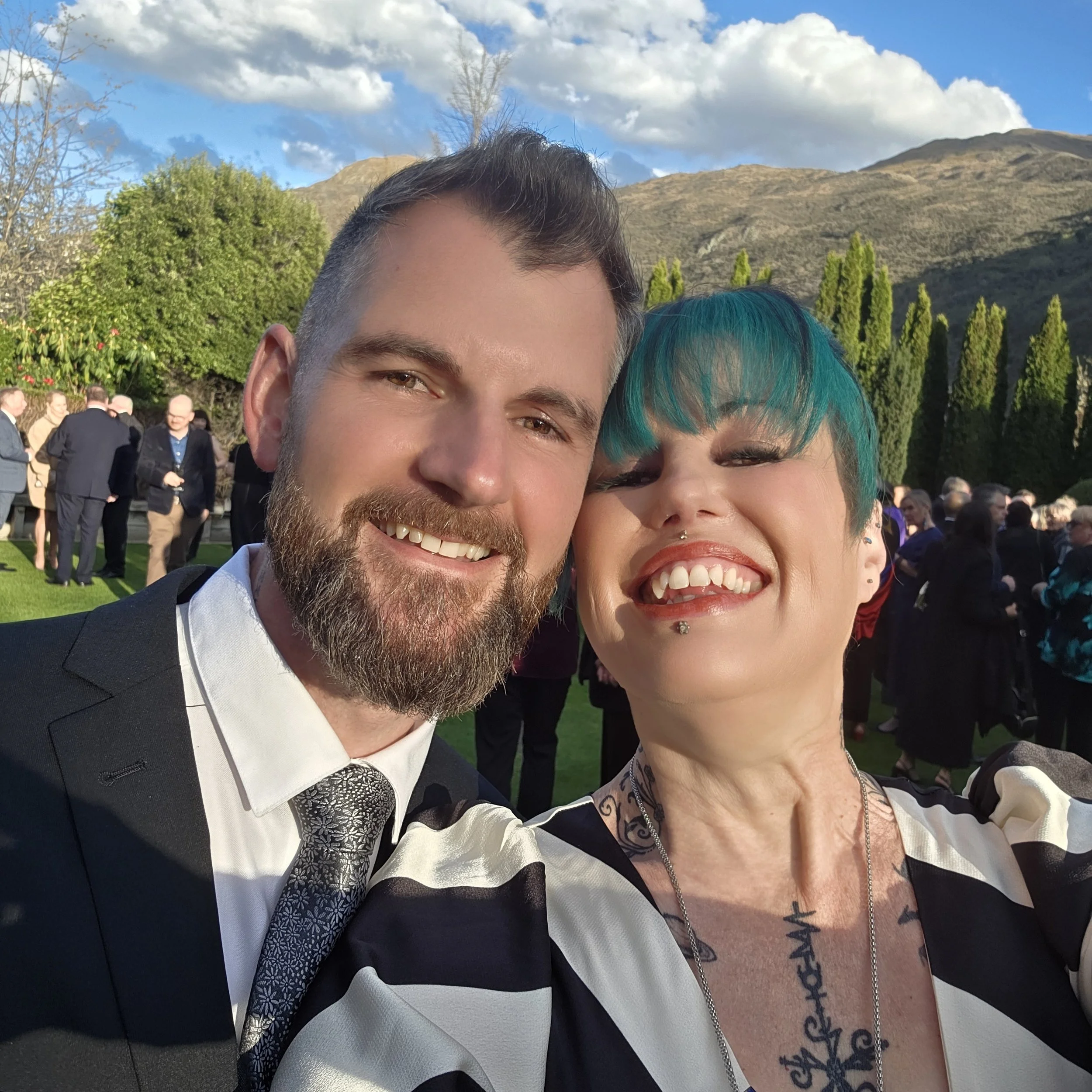 A man with a beard and a woman with teal hair smiling in an outdoor setting with mountains and trees in the background, at a gathering or event.