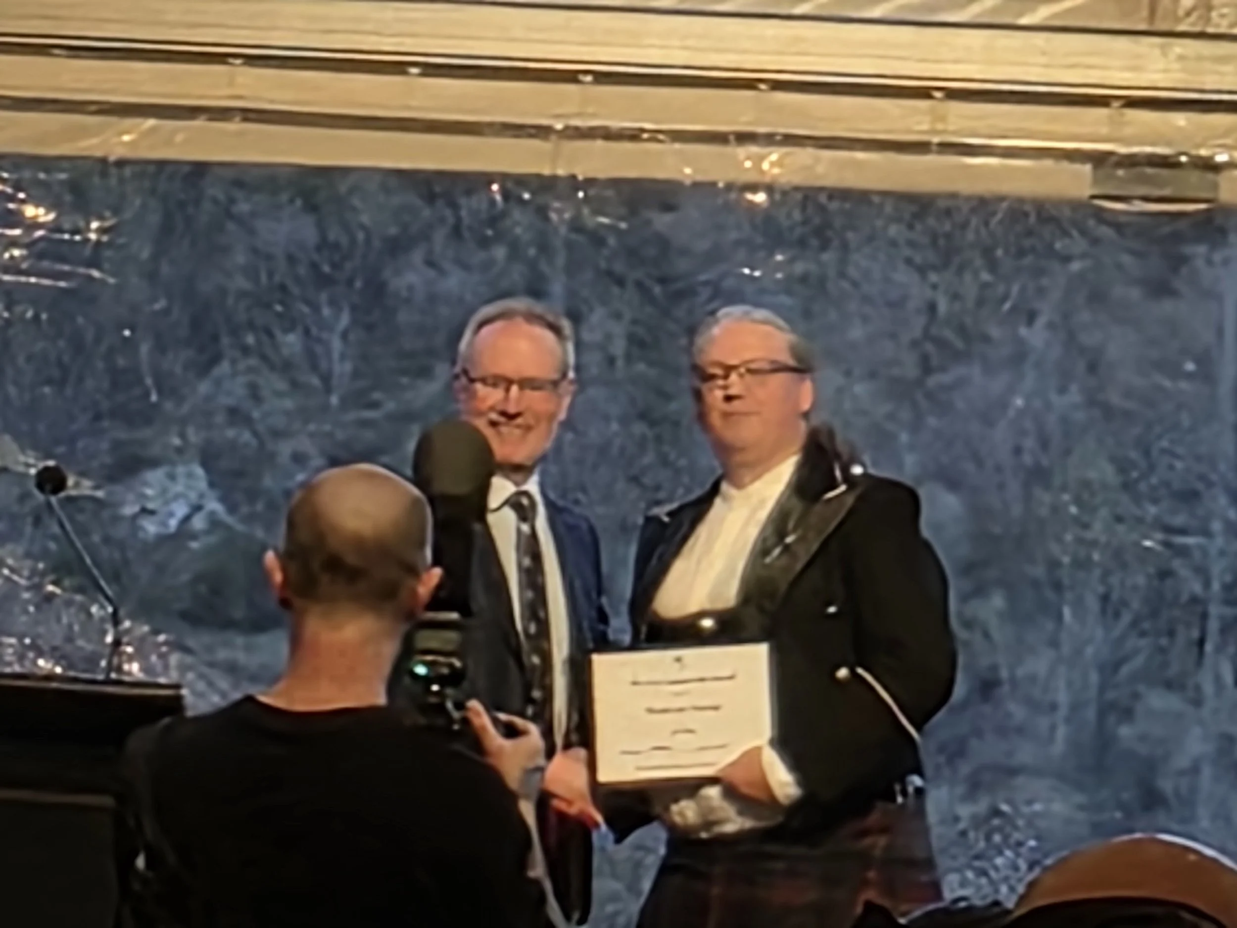 Two men on stage, one holding a certificate, posing for a photo as a photographer takes their picture. The background features a marble wall and a staircase.