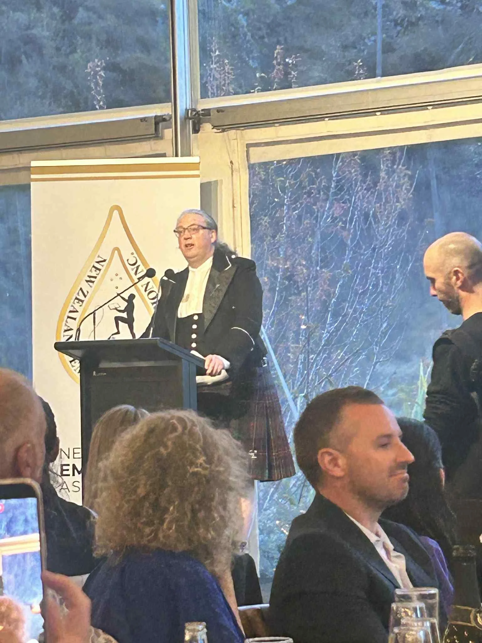 A man dressed in traditional Scottish attire, including a kilt, giving a speech at a podium during a formal event, with audience members seated in front of him.