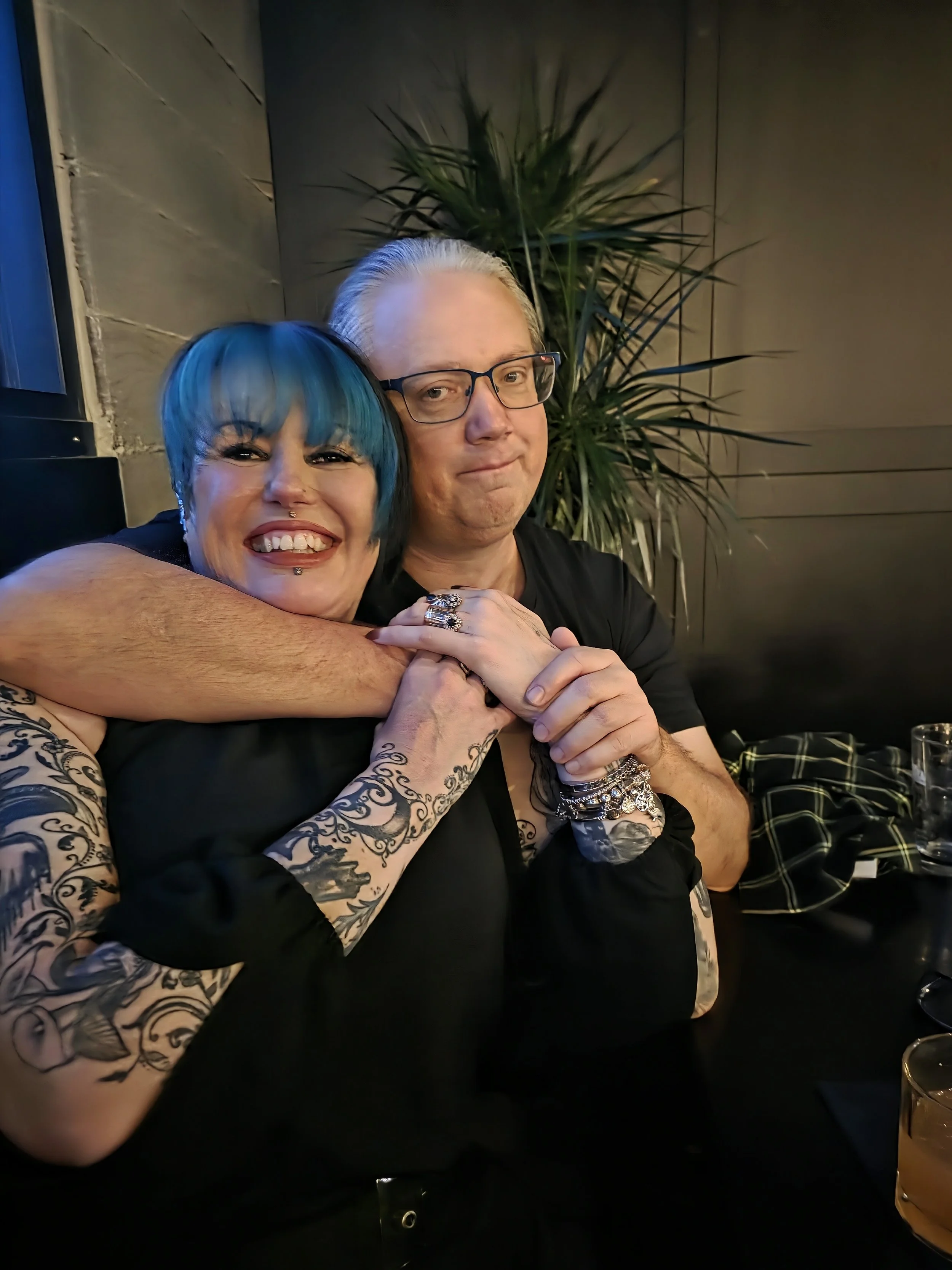 A woman with blue hair and tattoos smiling while hugging a man with glasses.