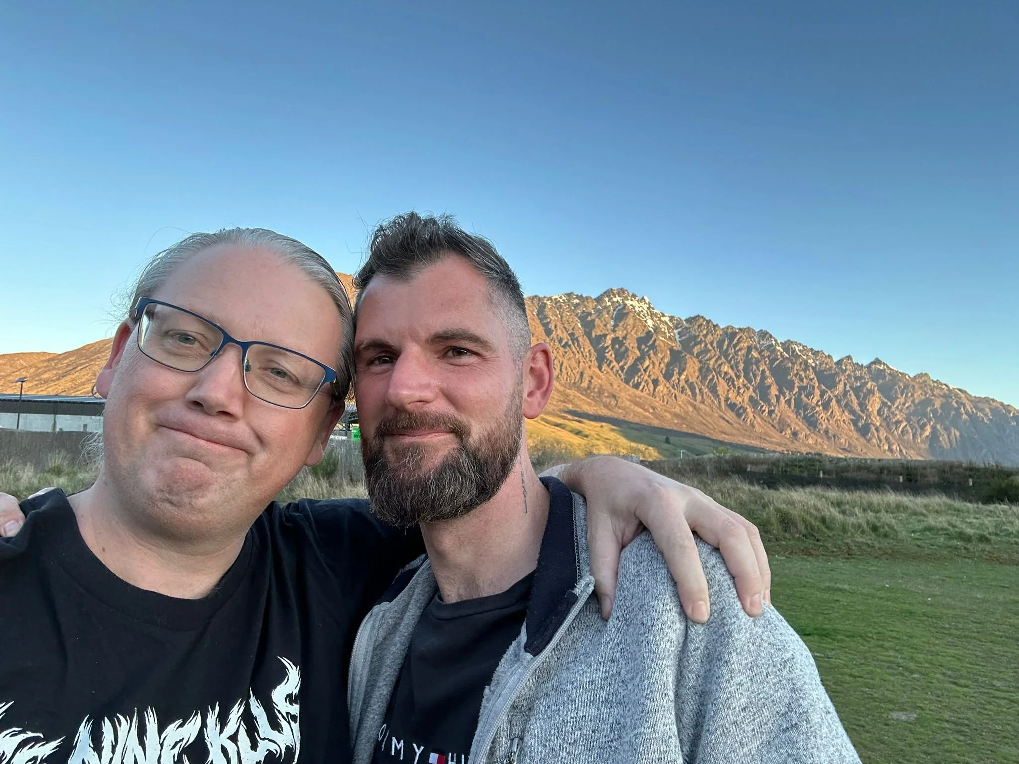 Two men taking a selfie with mountains in the background, one with glasses and the other with a beard, in an outdoor setting during sunset.