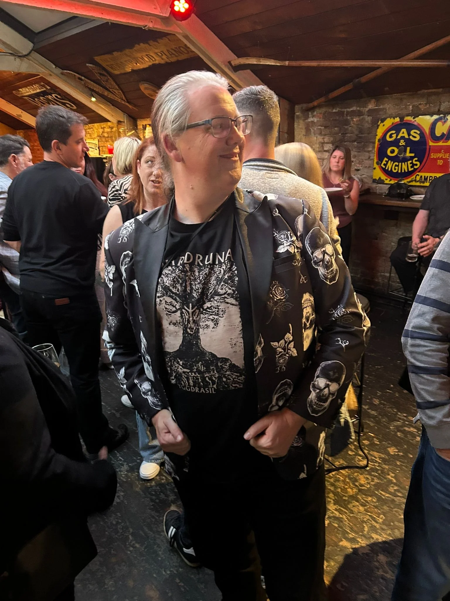 A man with glasses and gray hair, wearing a black jacket with skull and rose patterns, stands in a bar or pub filled with people, smiling and looking to the side.