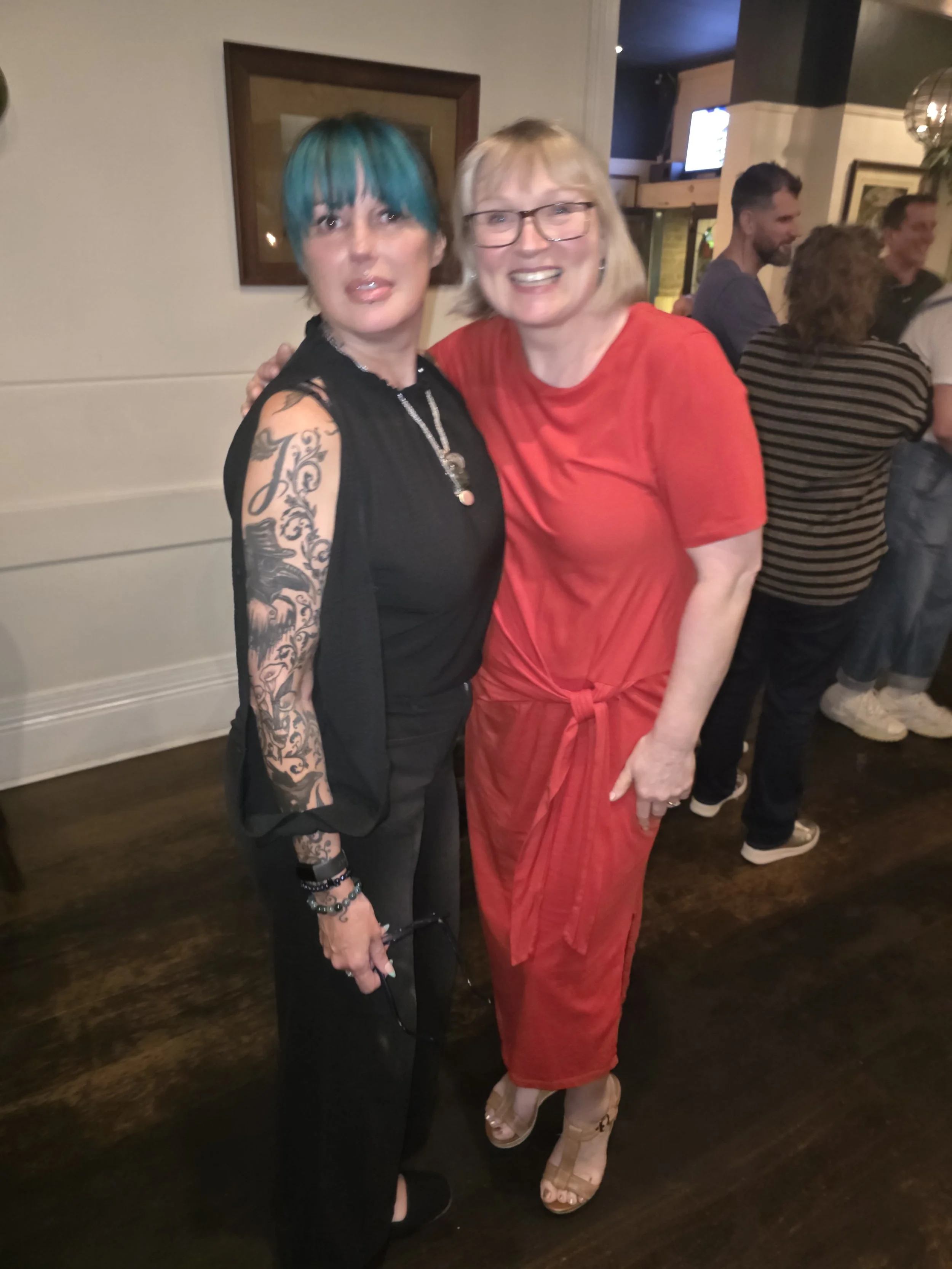 Two women standing close together and smiling indoors at a social gathering. The woman on the left has blue-tinted hair, tattoos on her right arm, and is wearing a sleeveless black top and black pants. The woman on the right has blonde hair, glasses,