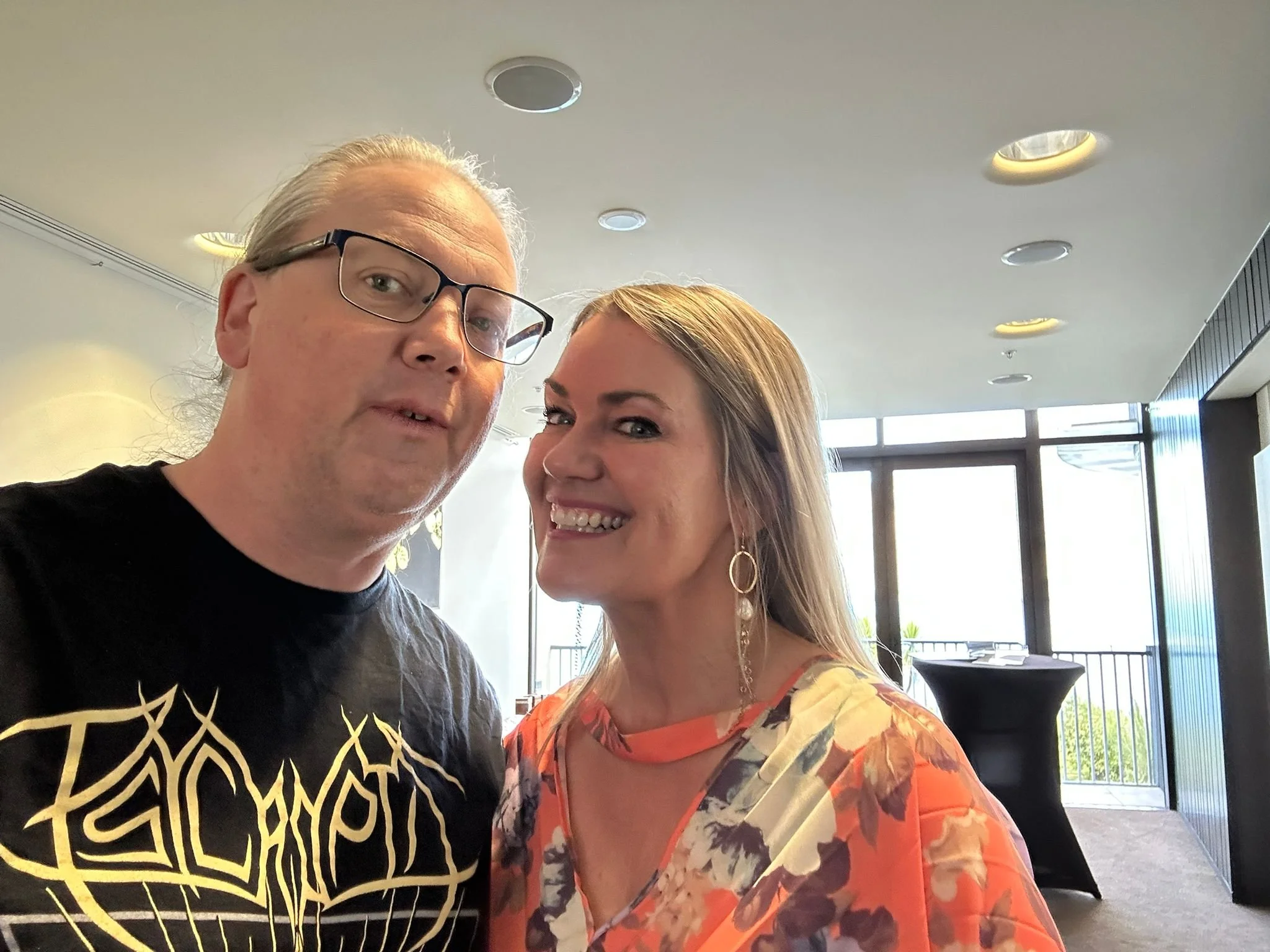 A man and a woman taking a selfie indoors during daytime, with large windows in the background letting in natural light.