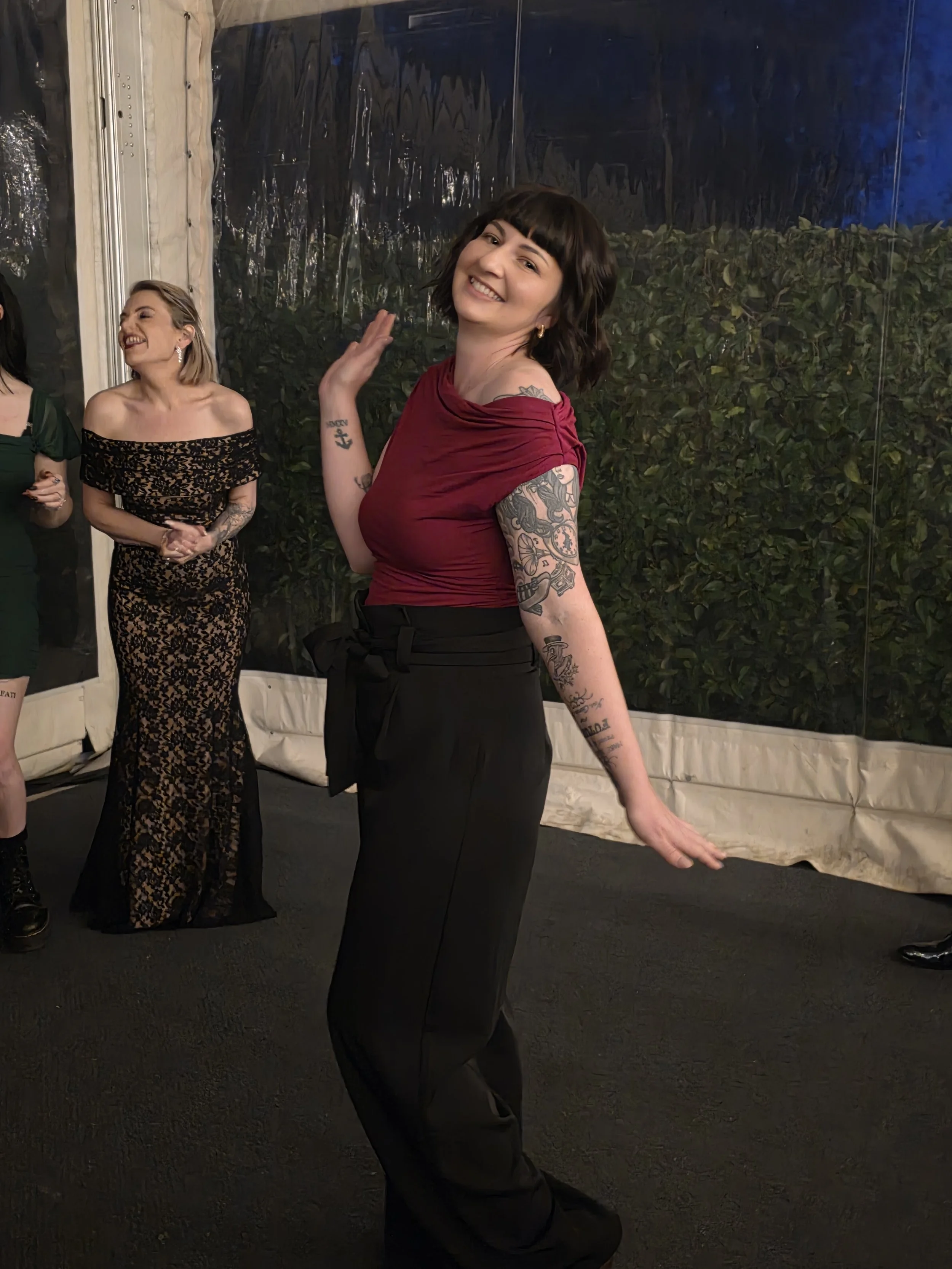 A woman with short dark hair and tattoos on her left arm, wearing a red top and black pants, dancing and smiling at an indoor event.