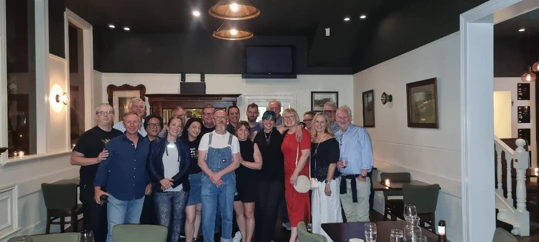 A group of about 20 people gathered in a restaurant with white walls and dark ceiling, some holding drinks, smiling for a group photo.