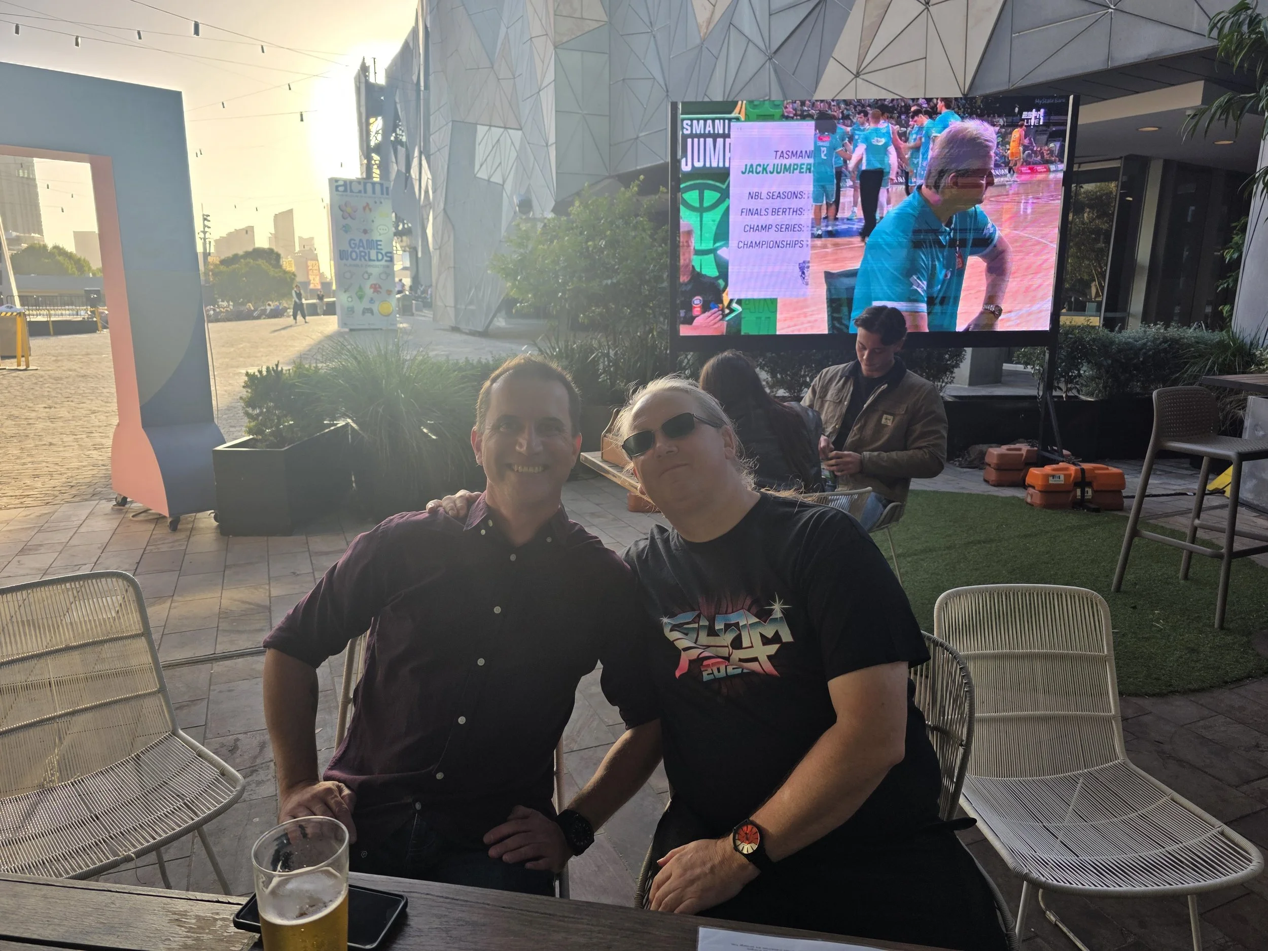 Two men sitting at an outdoor table, smiling, with a drink and a phone on the table. In the background, a large screen displays a sports event, and other people are seated nearby.