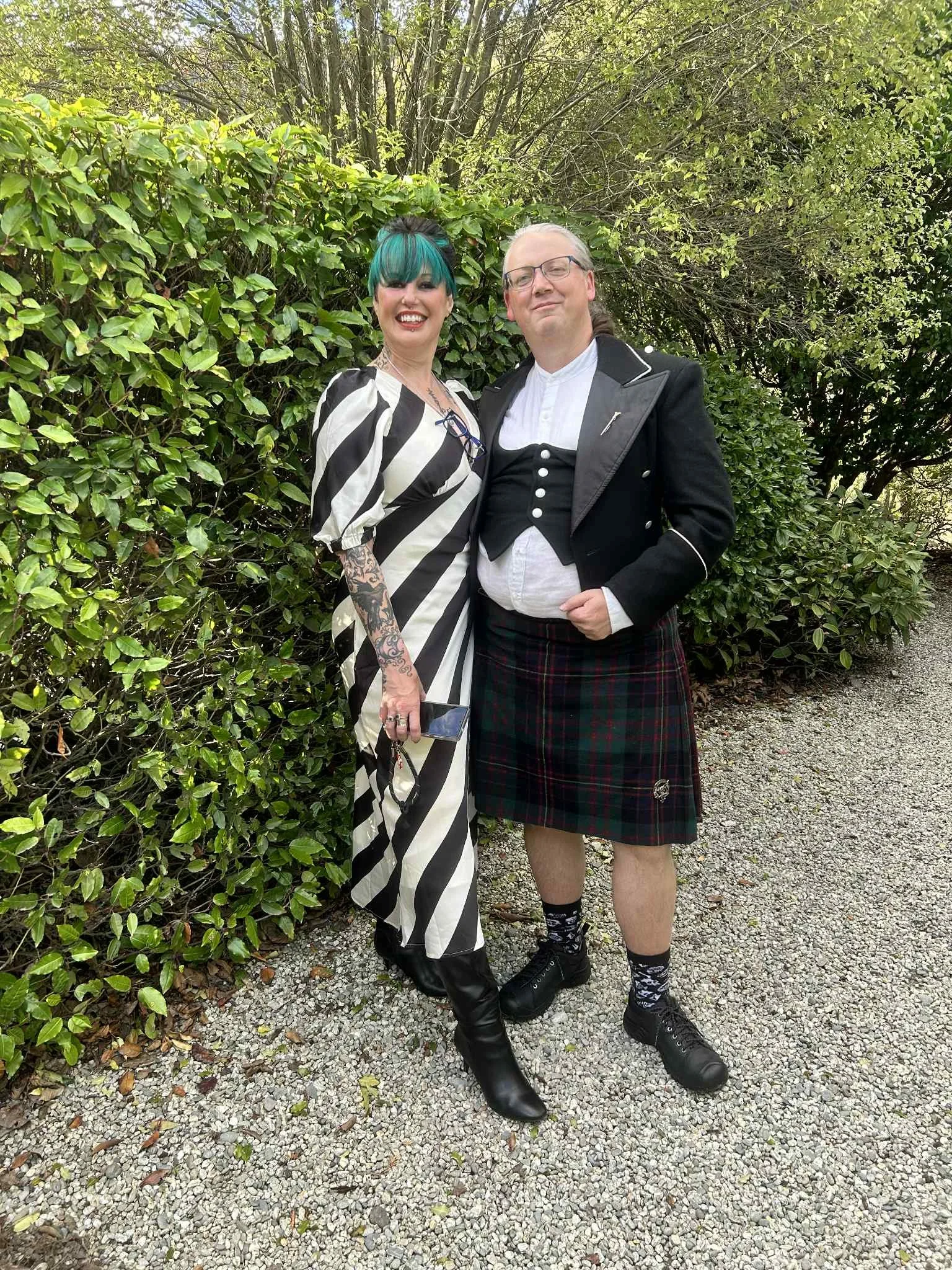 A woman with bright teal hair, in a black and white striped dress with tattoos on her arm, and a man dressed in traditional Scottish attire, including a kilt, jacket, and glasses, standing outdoors in front of green bushes and trees.