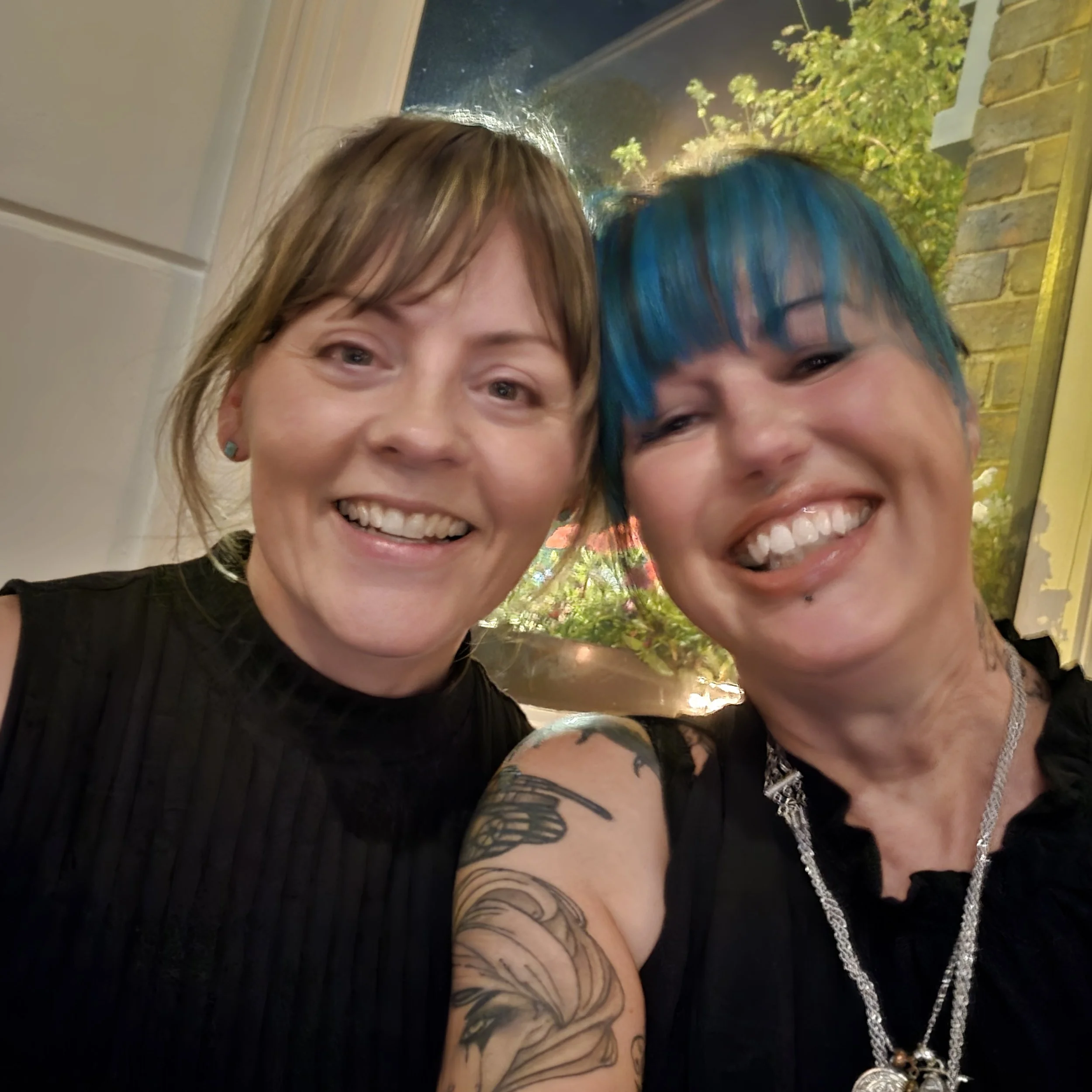 Two women smiling and taking a selfie, one with short brown hair and the other with short blue hair, both wearing black tops.