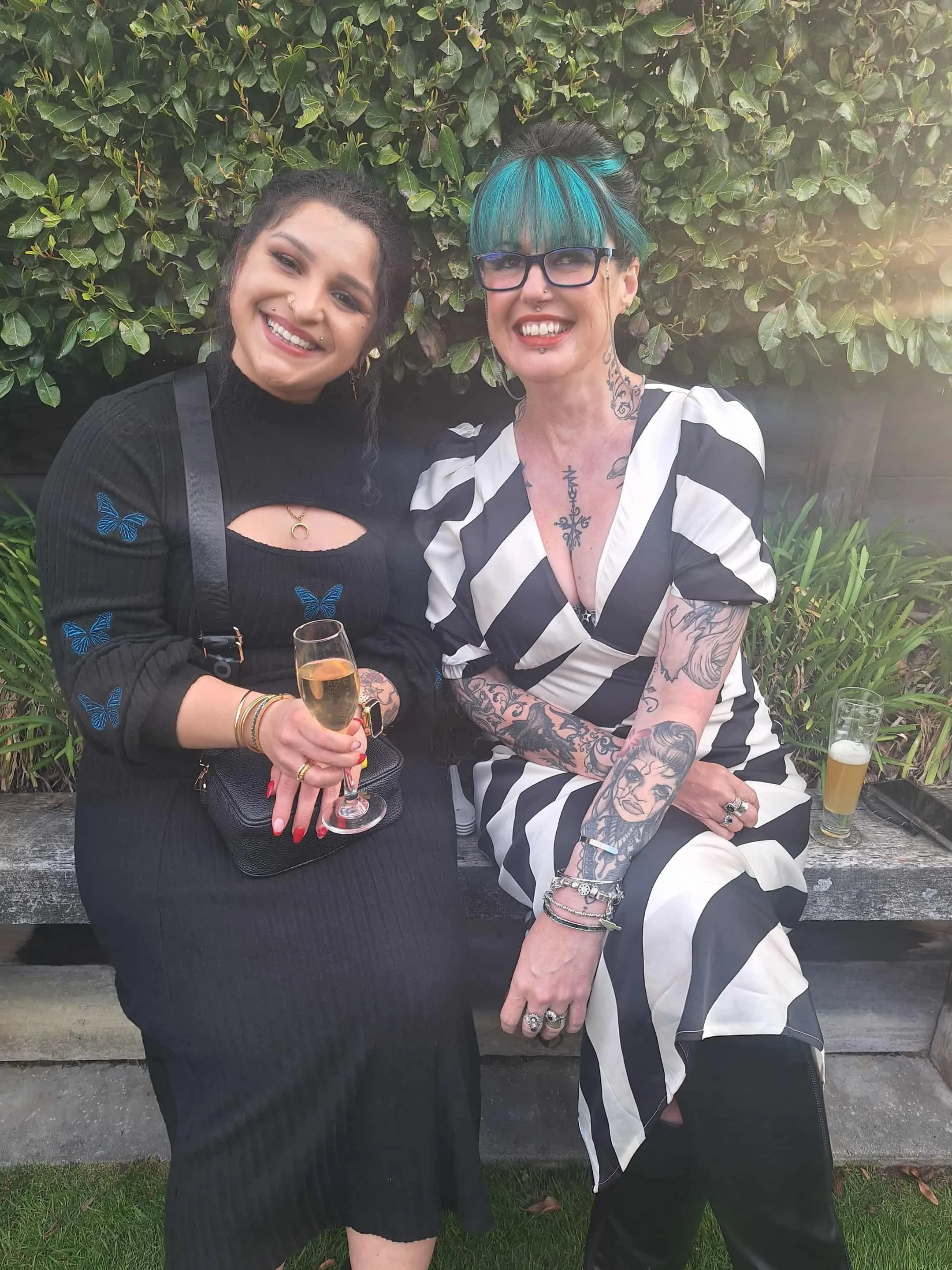 Two women sitting on a wooden bench in a garden, smiling and holding drinks, with greenery in the background.