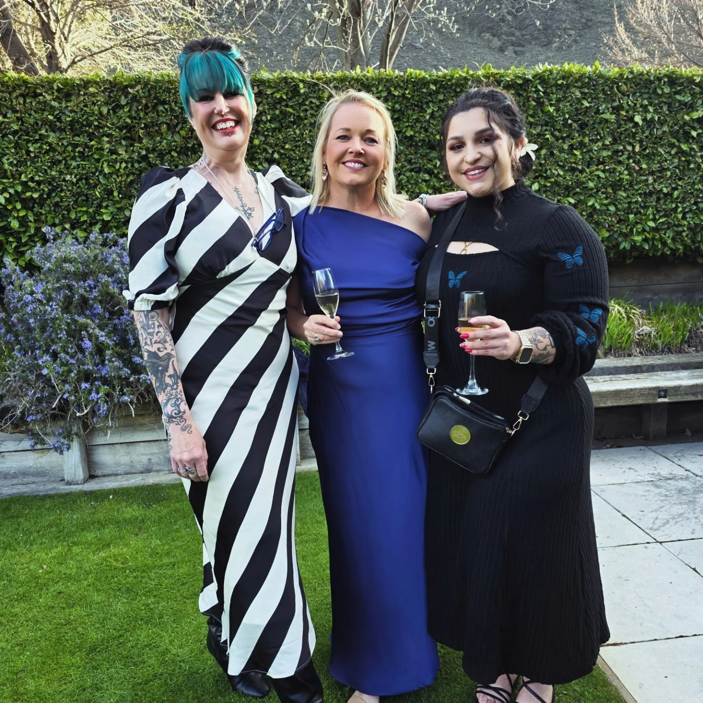 Three women standing outdoors, smiling and holding champagne glasses, with greenery and blooming plants in the background.