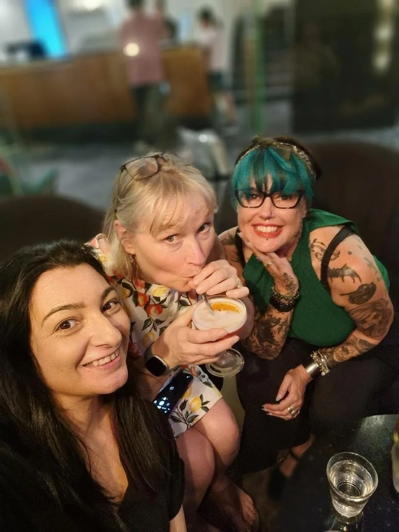 Three women sitting together at a bar or cafe, one is drinking a cocktail with a straw, all smiling and enjoying their time together.