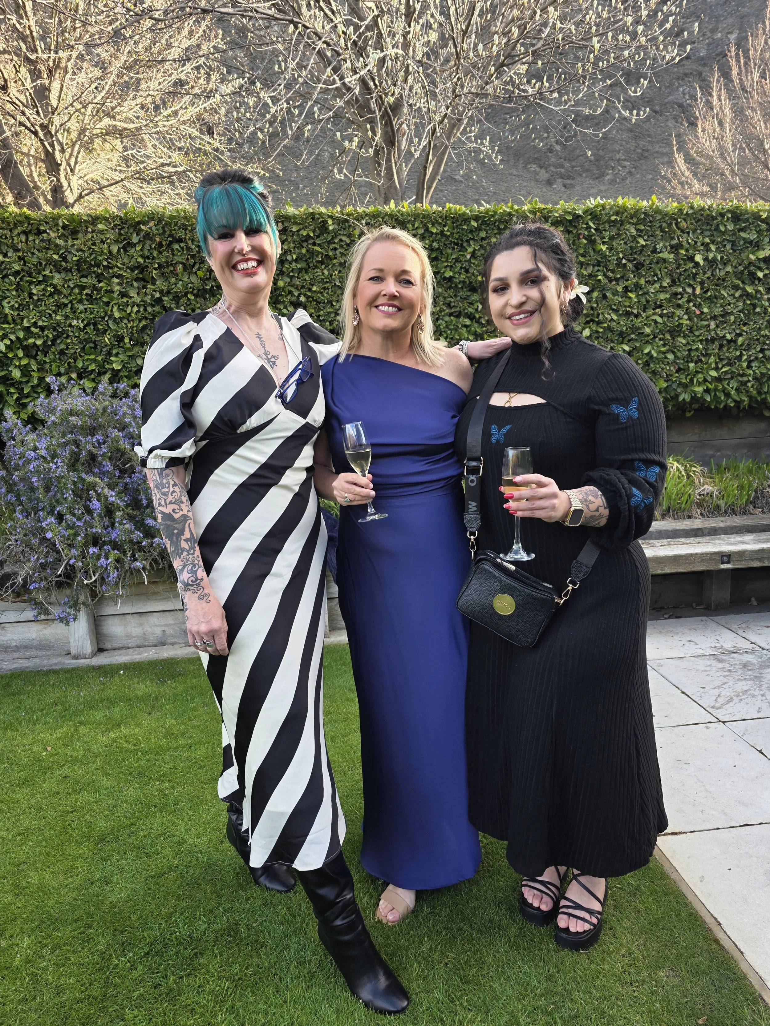 Three women standing outdoors on a lawn, dressed elegantly and holding glasses of champagne, smiling for the photo.