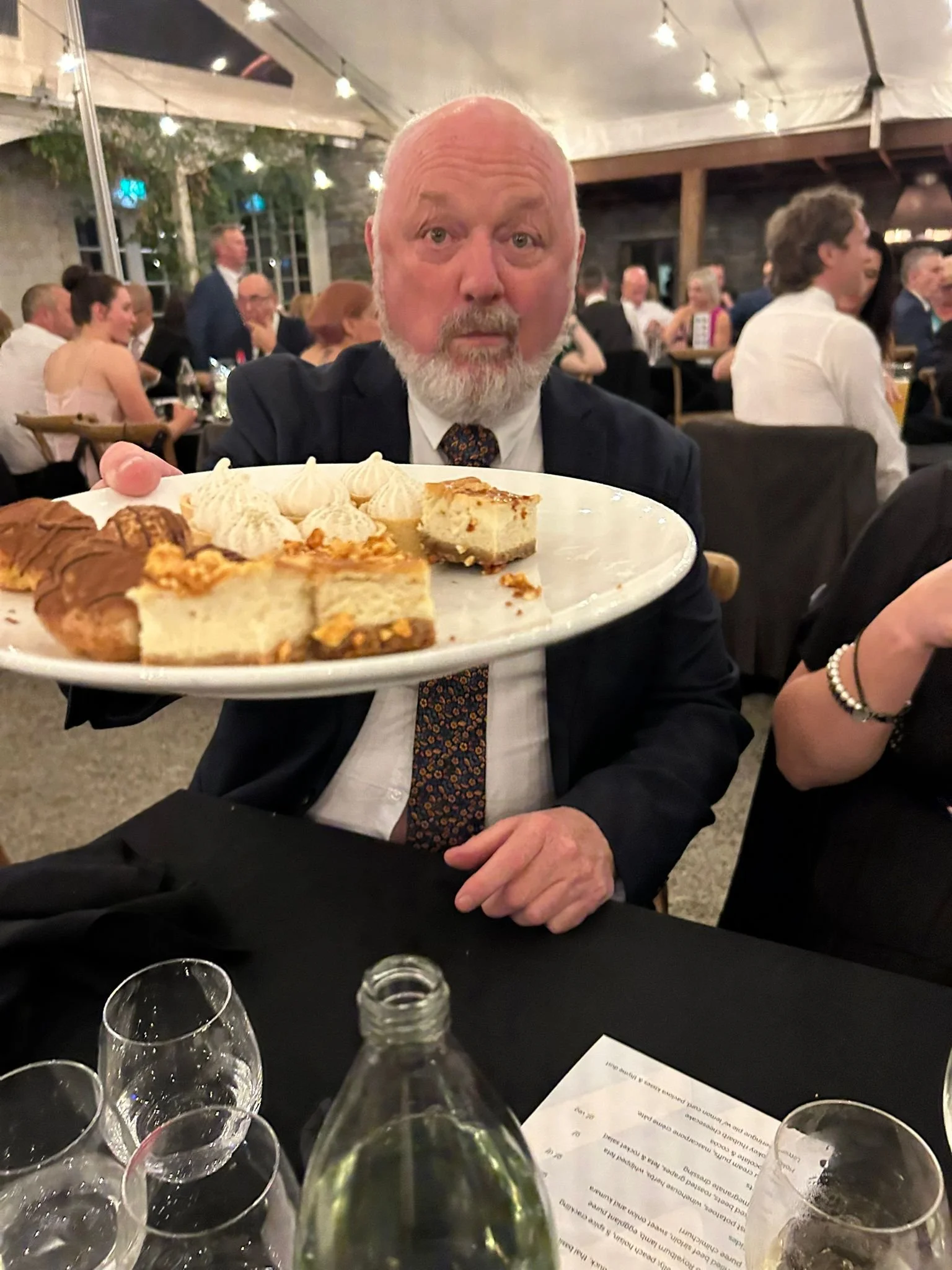 An older man with a beard, dressed in a dark suit, holding a large tray of assorted desserts at a social event.