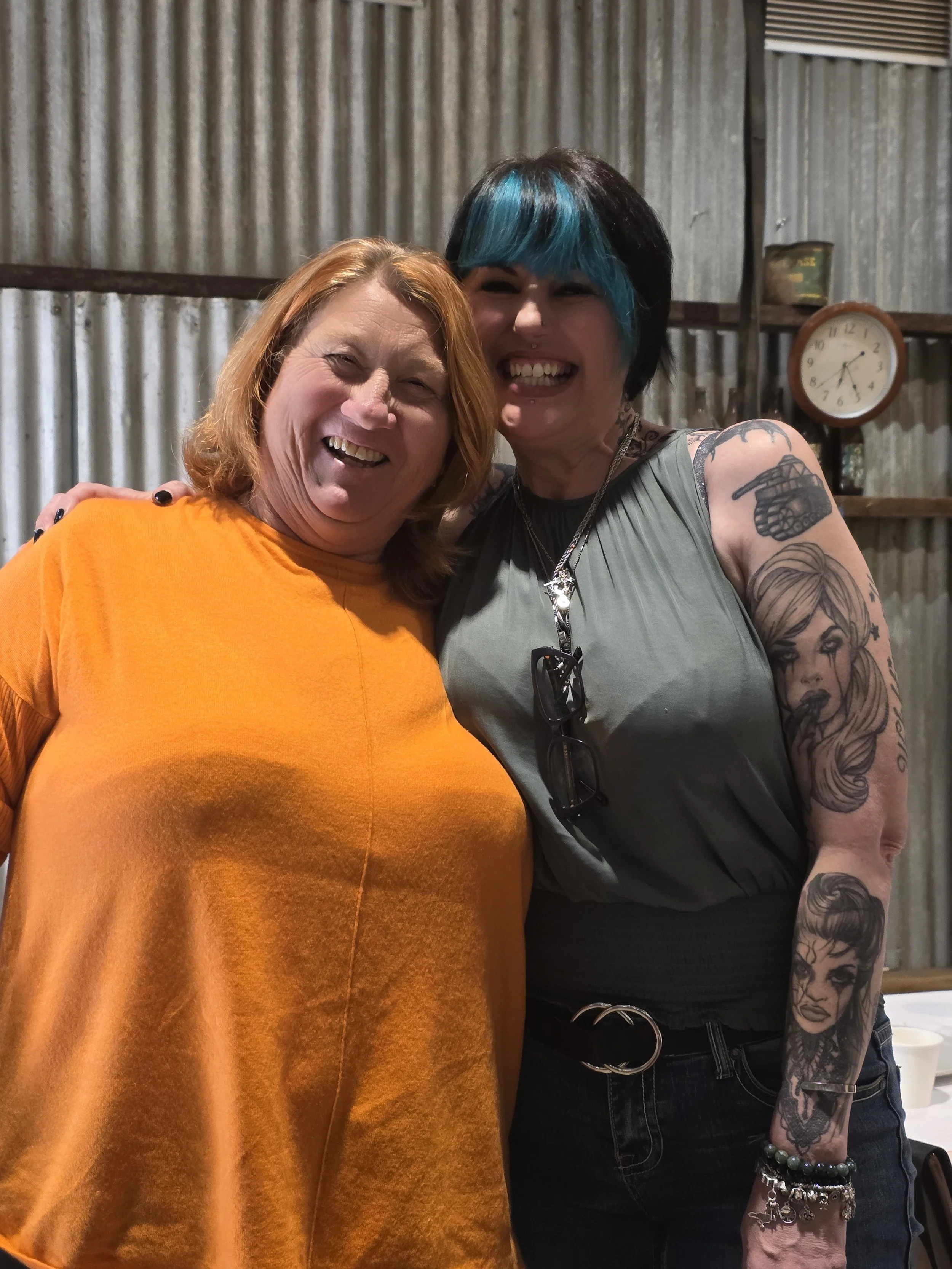 Two women smiling and hugging in front of a rustic metal wall, one with red hair in an orange shirt, and the other with black hair with turquoise streaks in a sleeveless top, tattoos on her arms, wearing glasses hanging from her shirt, and bracelets.