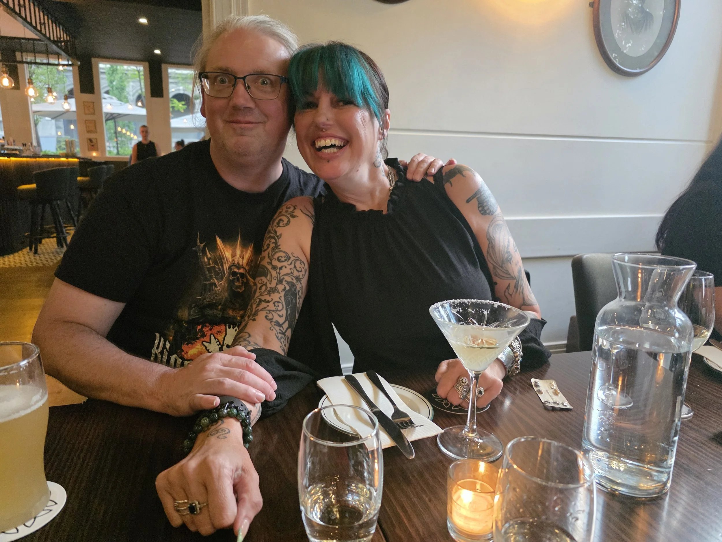 A man and woman sitting at a restaurant table, smiling and posing for a photo. The man has light hair, glasses, and wears a black t-shirt. The woman has short hair with teal streaks, tattoos on her arms, and wears a sleeveless black top. There are dr