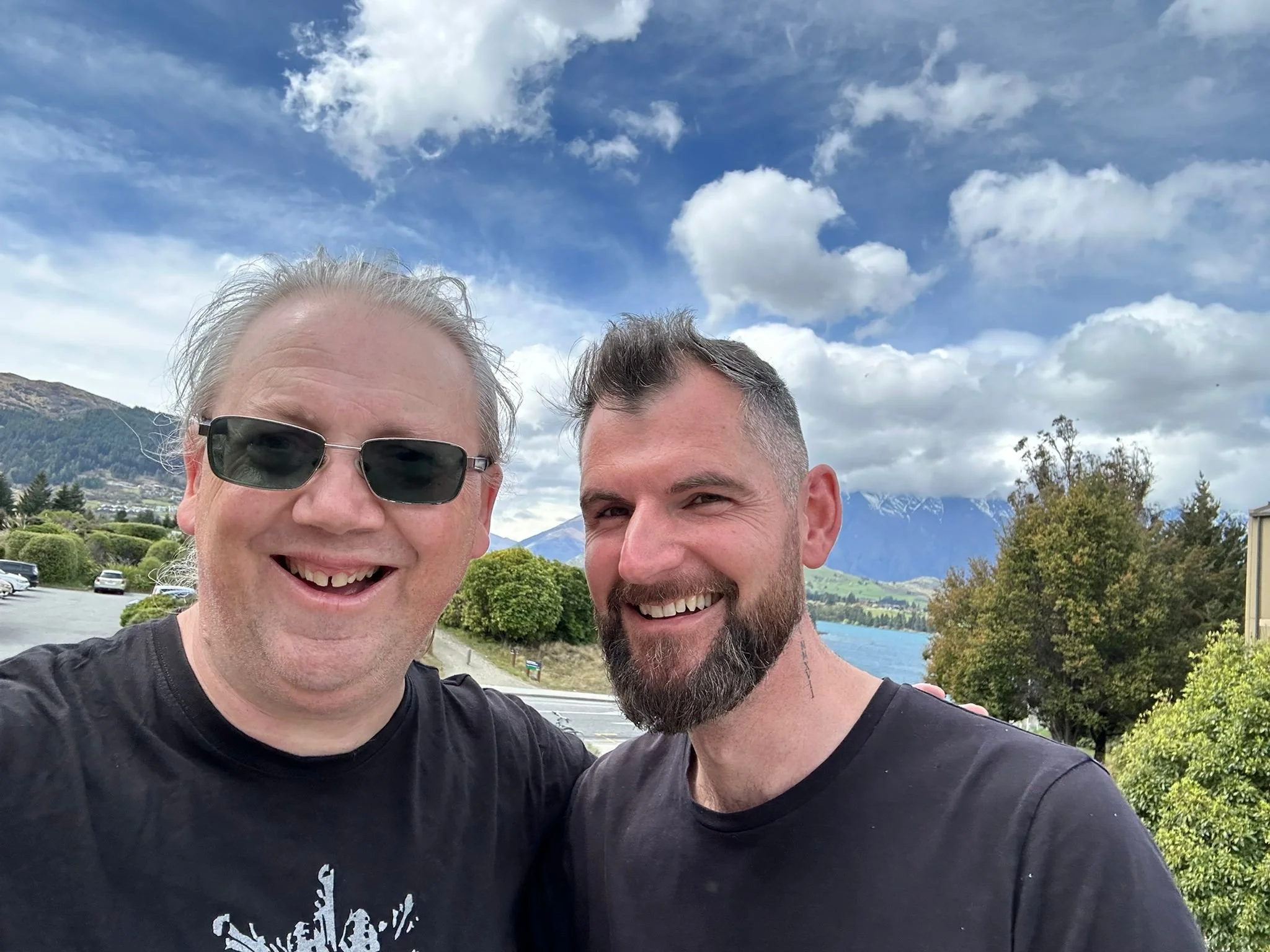 Two smiling men standing outdoors with a scenic lake, mountains, and trees in the background.