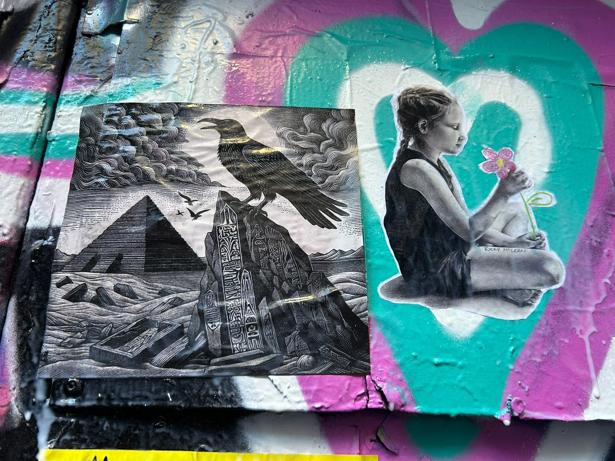 Street art graffiti featuring a woman sitting cross-legged, coloring a flower, and a monochrome print of a raven above pyramids with a stormy sky in the background. The artwork has vibrant pink, teal, and purple colors.