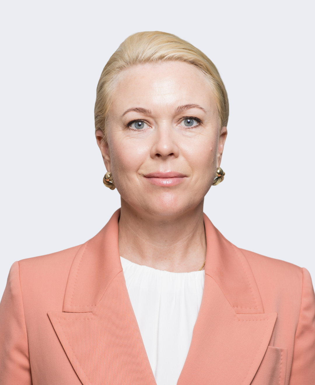 A professional woman with blonde hair, blue eyes, and fair skin, wearing a peach blazer and white blouse, against a plain light gray background.