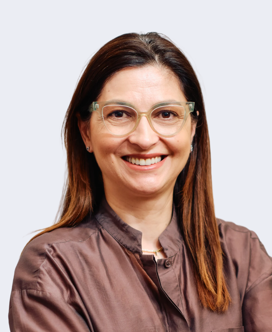 A woman with brown hair and glasses smiling at the camera, wearing a brown blouse, against a light gray background.