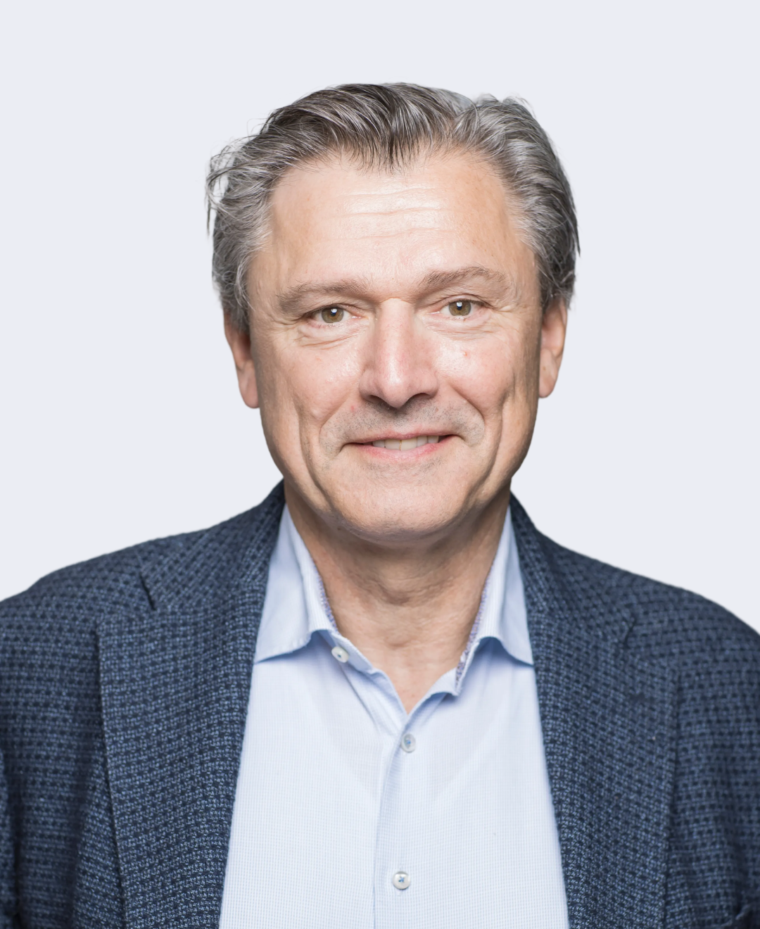 A portrait of a middle-aged man with gray hair, wearing a blue blazer and a light blue shirt, smiling softly against a plain light background.