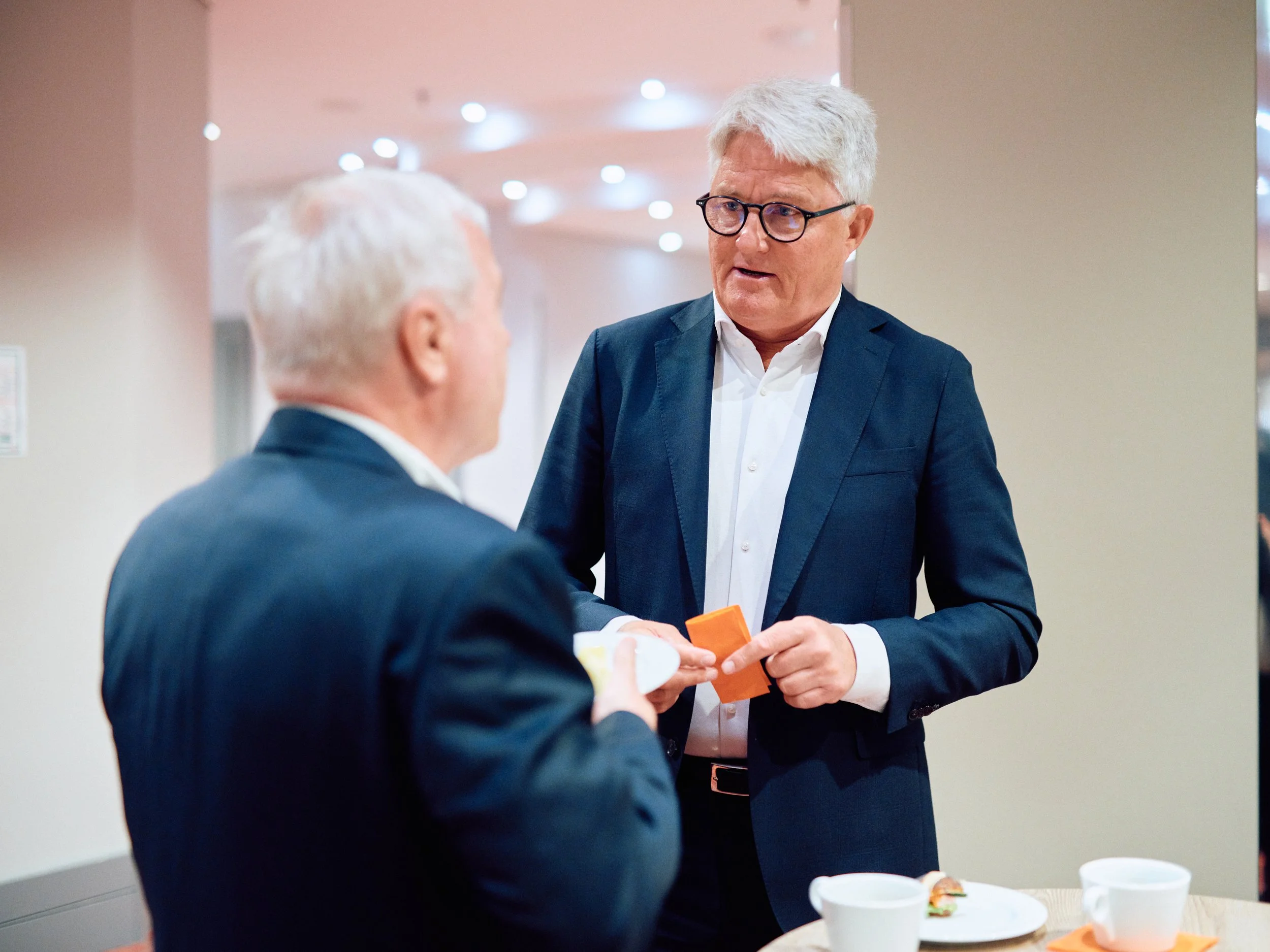 Two men in suits shaking hands and exchanging an orange envelope at a professional event or reception, with food and drinks on the table in the foreground and a softly lit background.