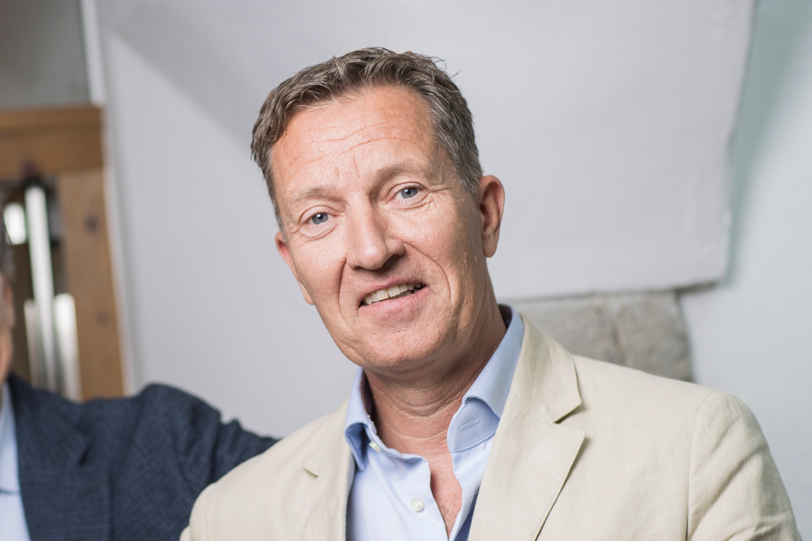 A middle-aged man with light brown hair and blue eyes wearing a light beige blazer over a blue collared shirt, smiling slightly in an indoor setting.