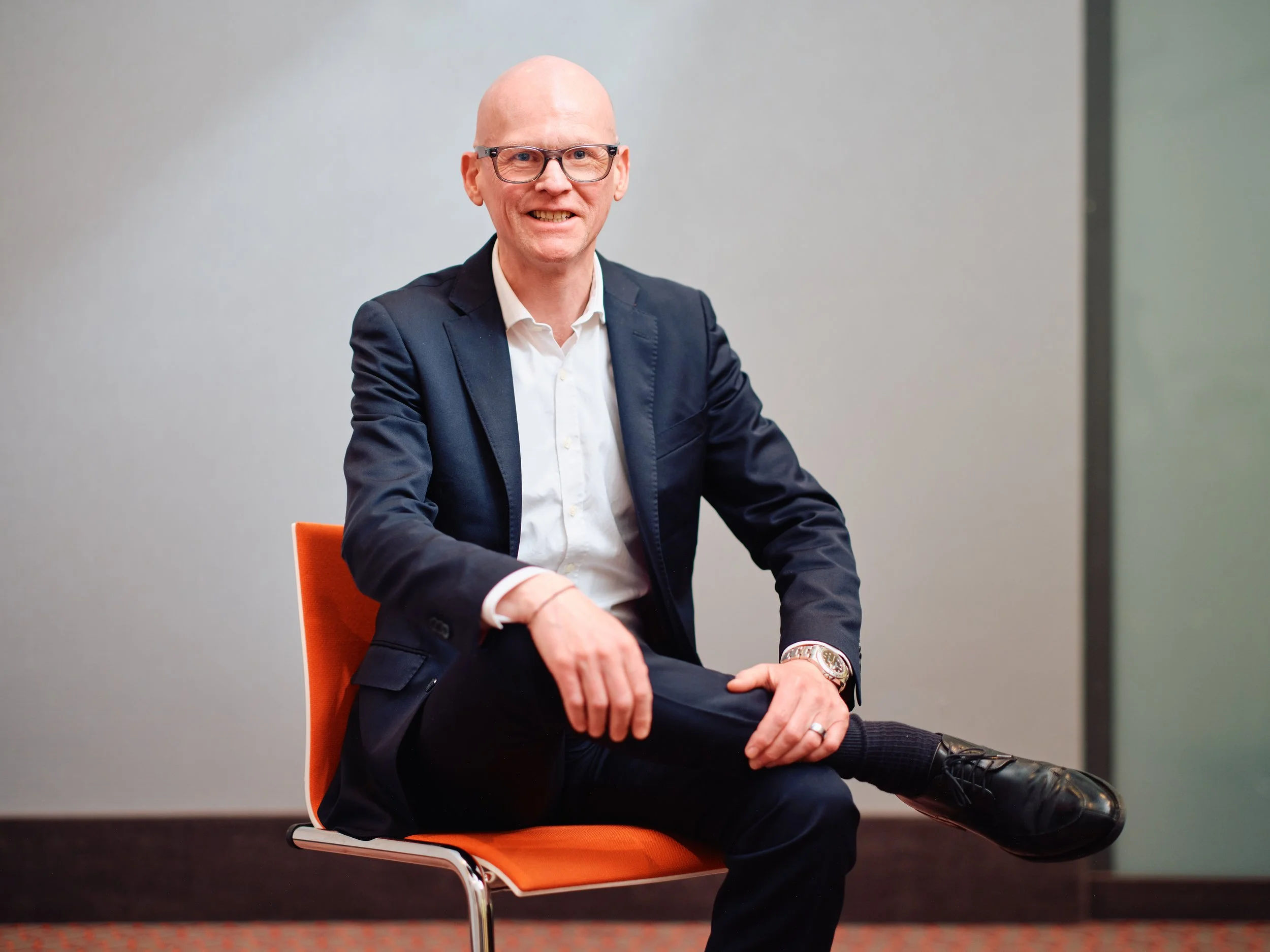 A bald man with glasses, wearing a dark blazer and white shirt, sitting on an orange chair in front of a plain, gray wall, smiling at the camera.