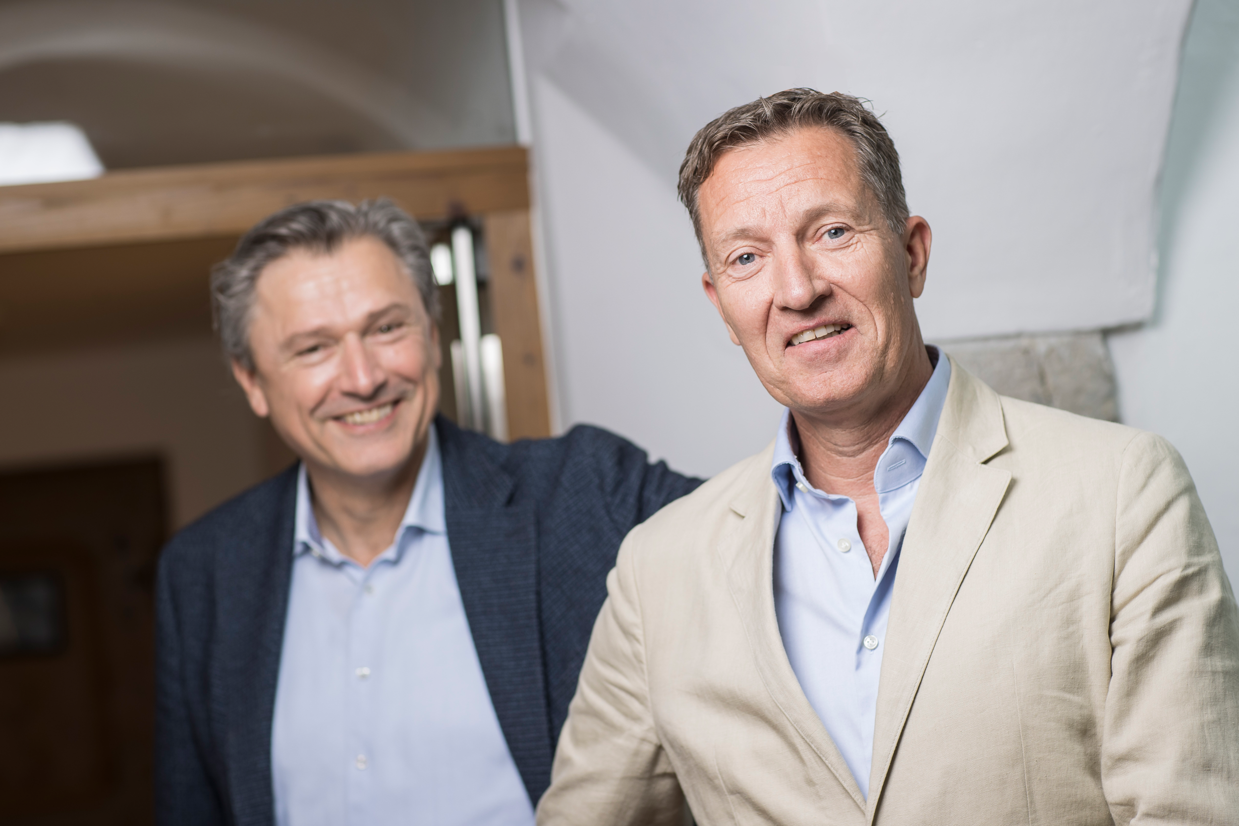 Two middle-aged men smiling and posing for a photo, one in a beige blazer and the other in a dark blazer, in an indoor setting.