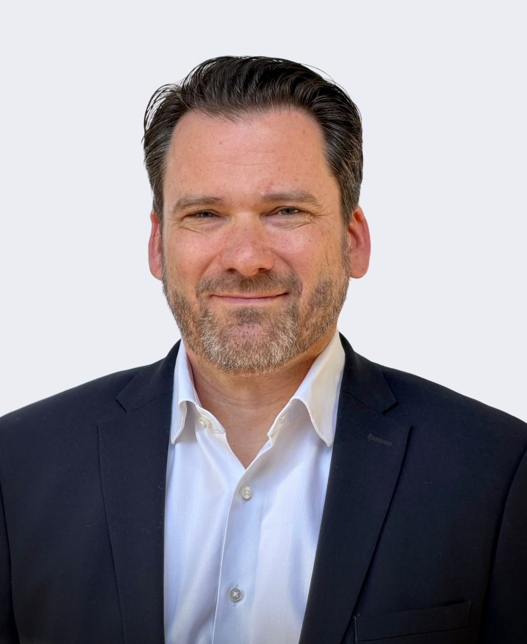 A middle-aged man with dark hair, a beard, and a slight smile, wearing a white collared shirt and a dark blazer, posing against a plain light-colored background.