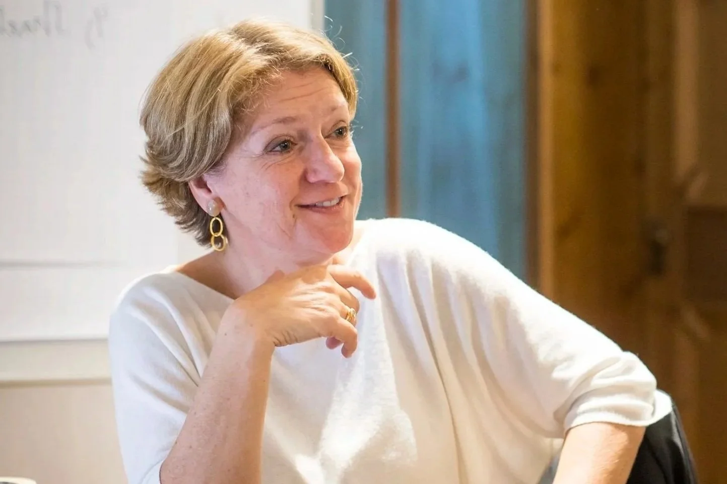 A woman with short blond hair, wearing gold earrings and a white top, smiling and resting her chin on her hand in a room with wooden furniture.