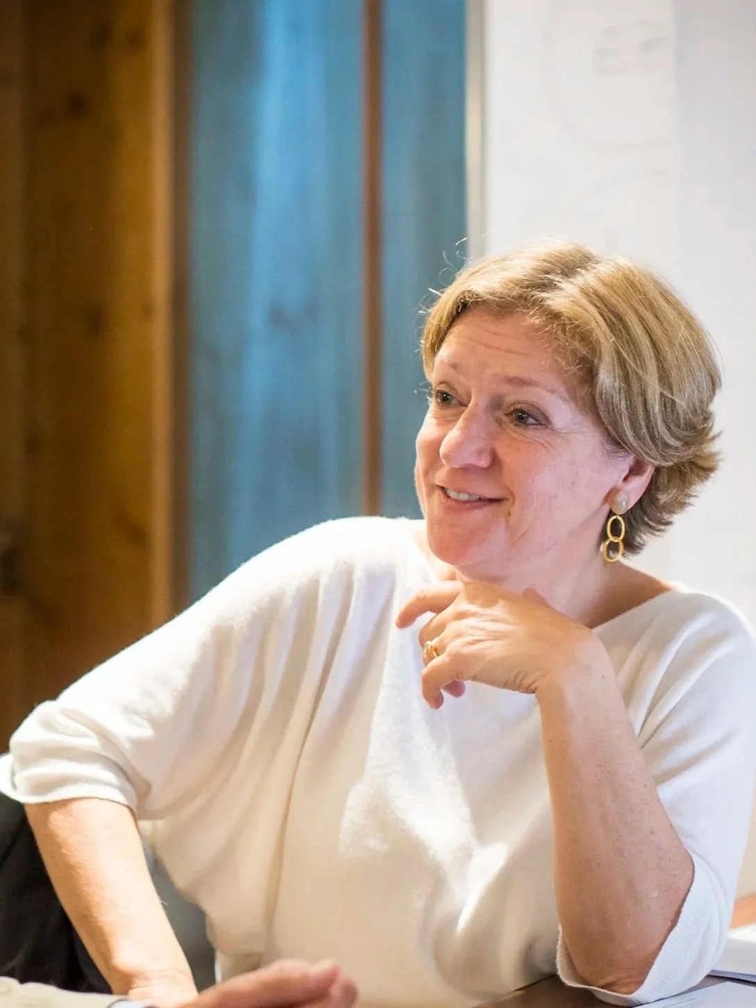 A woman with short blond hair, wearing gold earrings and a white top, smiling and resting her chin on her hand.