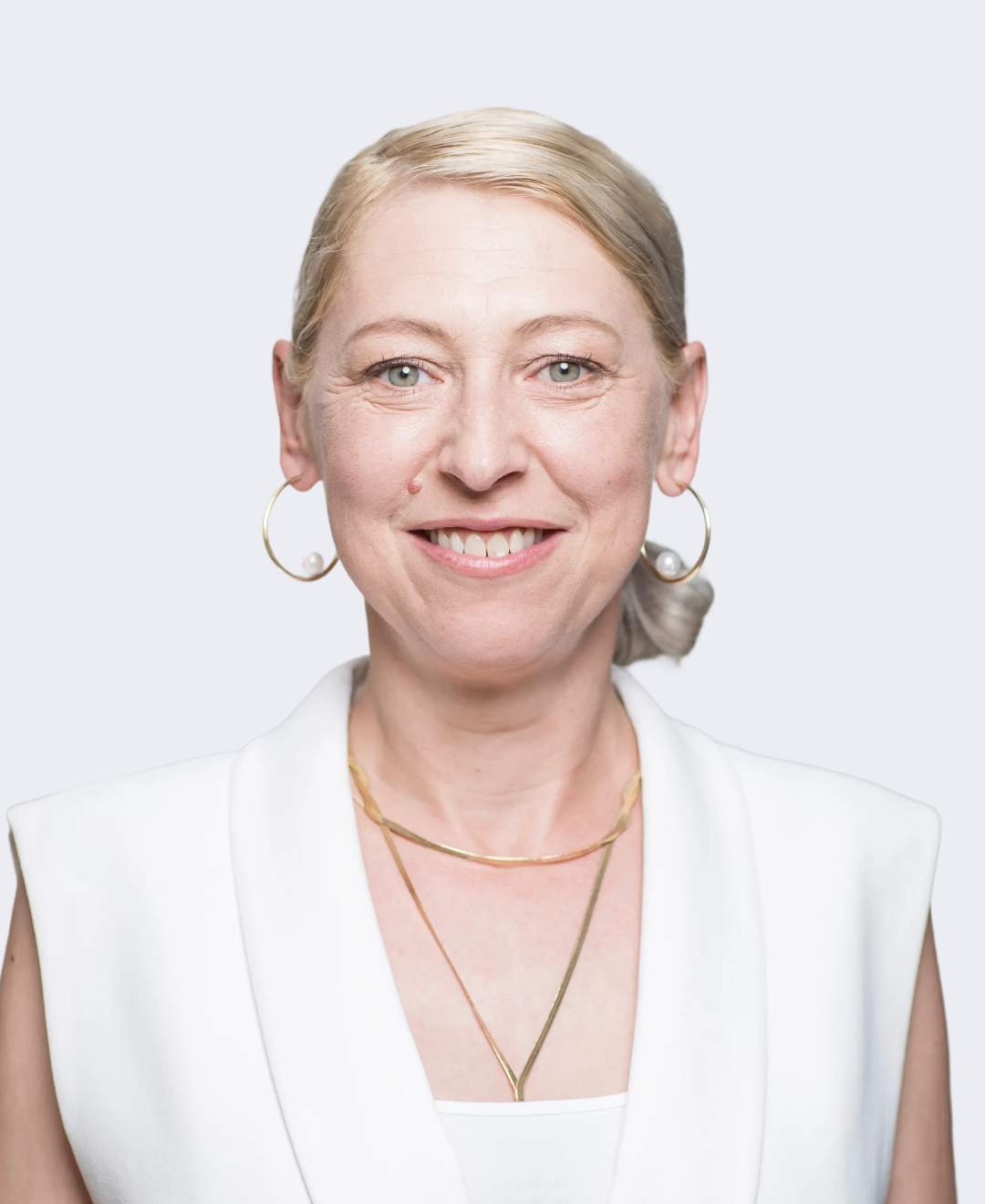 Portrait of a smiling woman with blonde hair, wearing hoop earrings and layered gold necklaces, dressed in white against a light background.