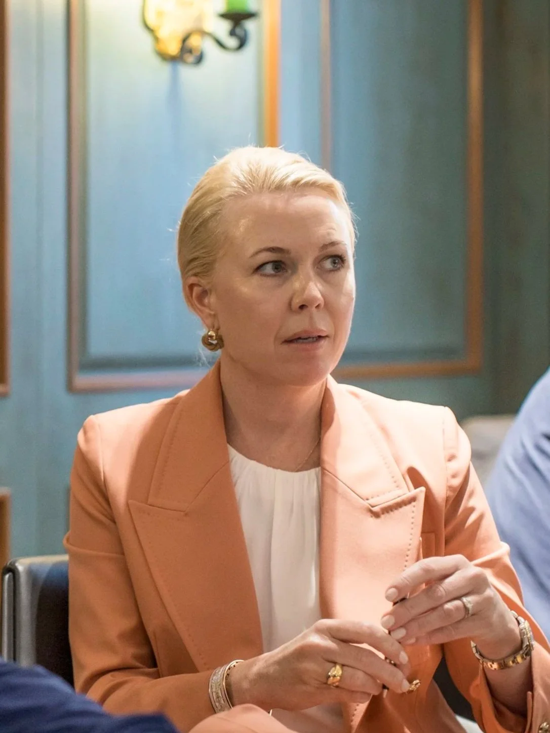 A woman with blonde hair in an updo, wearing a peach blazer and white shirt, sitting at a table, holding a glass, with a blue and gold decorative wall behind her.