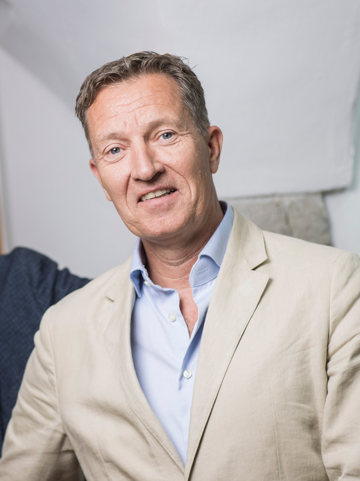 A middle-aged man with short, light brown hair and blue eyes, wearing a beige blazer and light blue shirt, smiling at the camera. The background features a white wall and part of a stone fireplace.